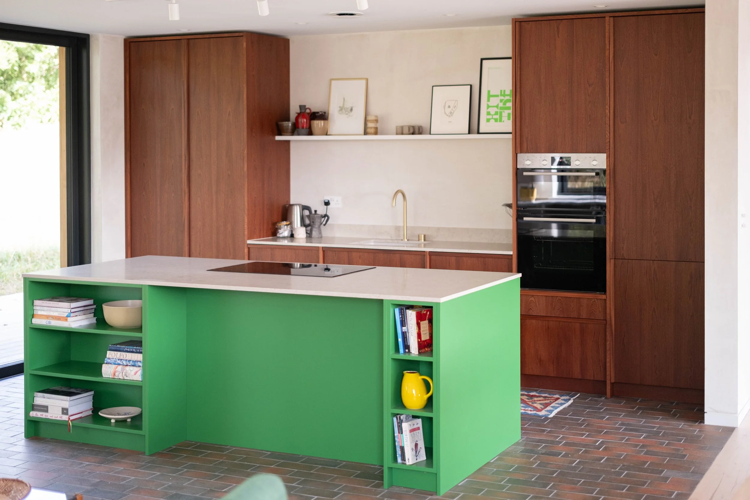 A modern kitchen featuring dark wood cabinets, a white countertop with a gold faucet, and a bright green island with books and a yellow vase. Art prints and decorative bowls are on a white shelf above the countertop, and a large window or sliding doo