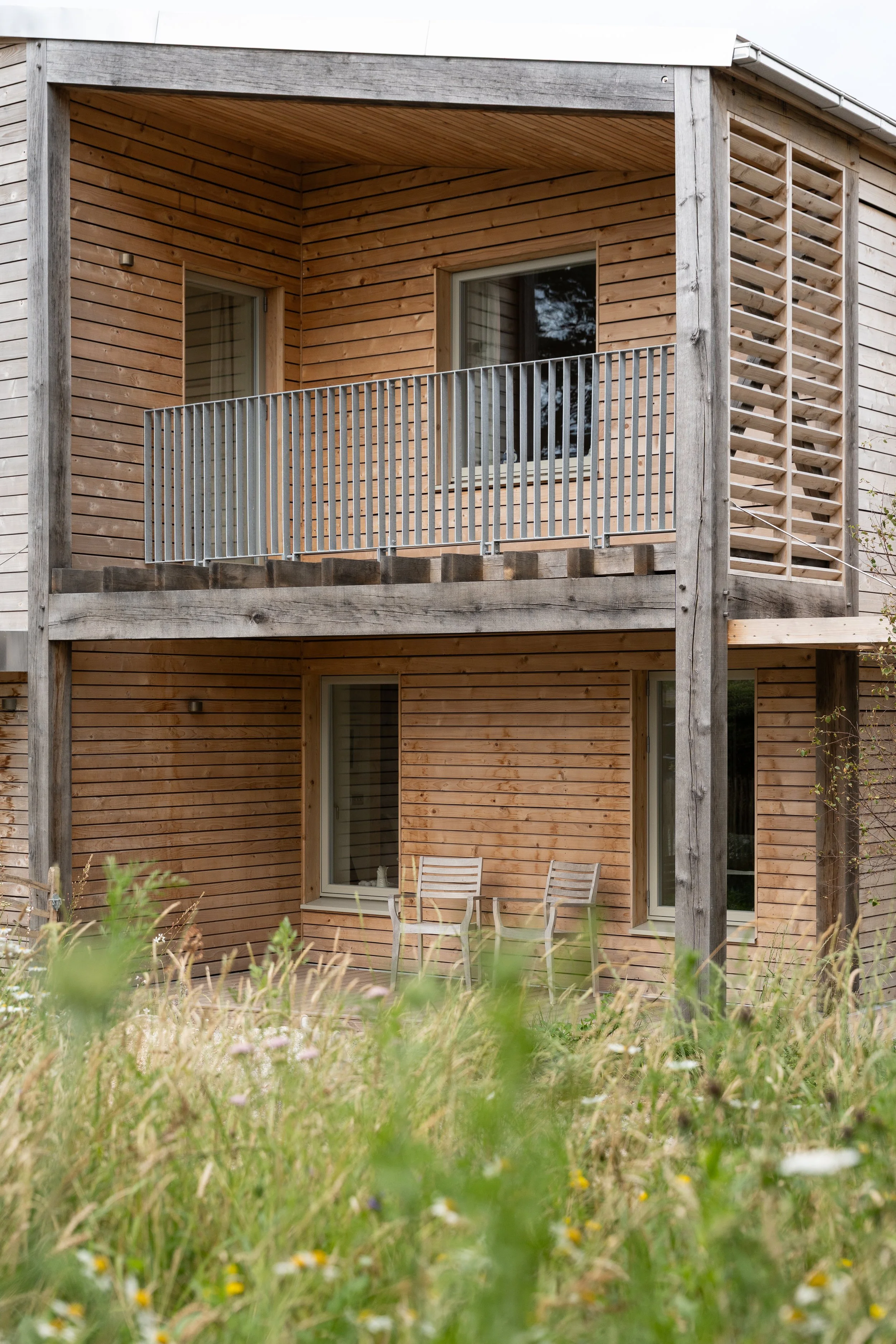 View of a wooden apartment building with a balcony and patio furniture in a grassy field.