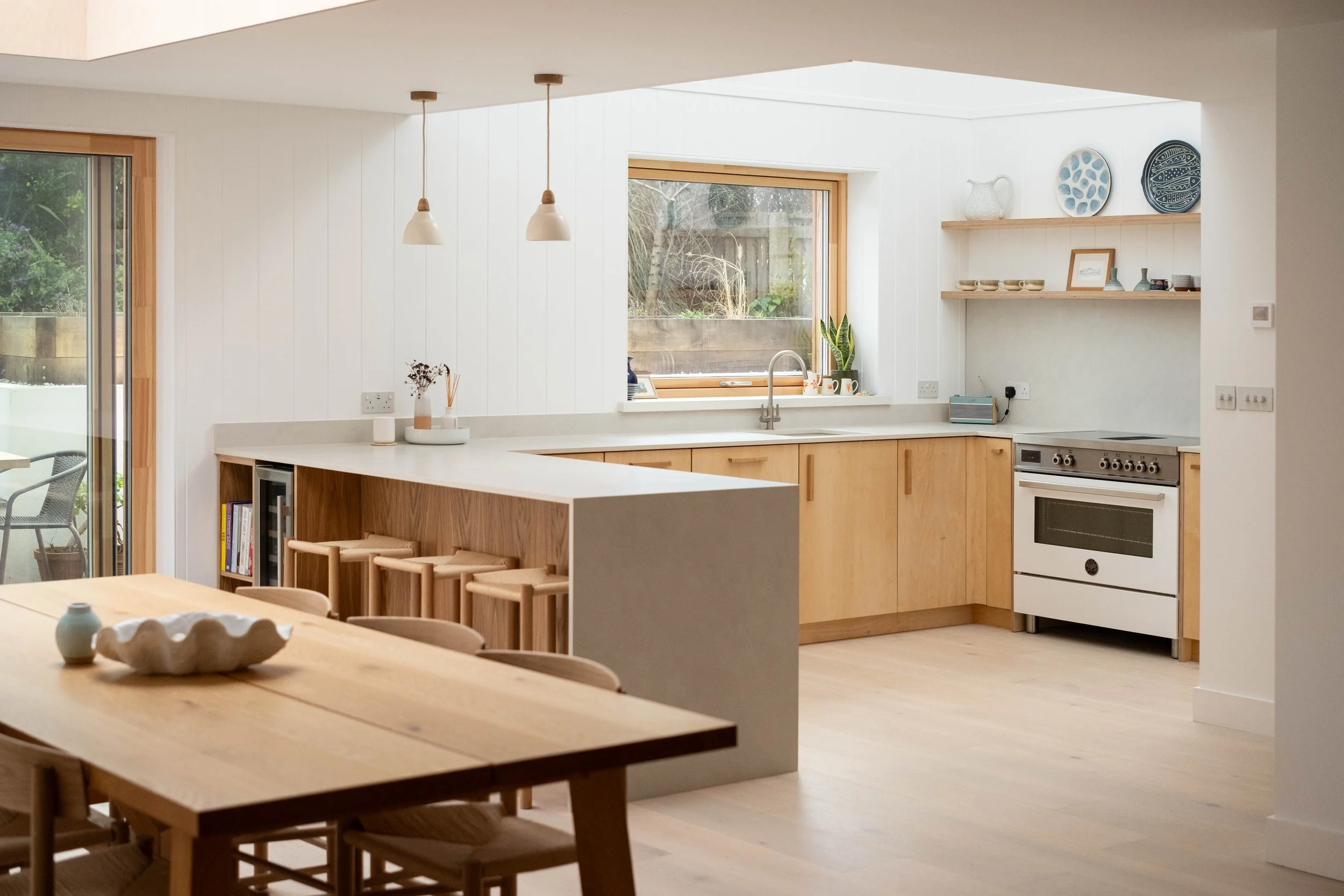 Kitchen with wooden cabinets, white countertops, large window, hanging pendant lights, and dining area with wooden table and chairs.