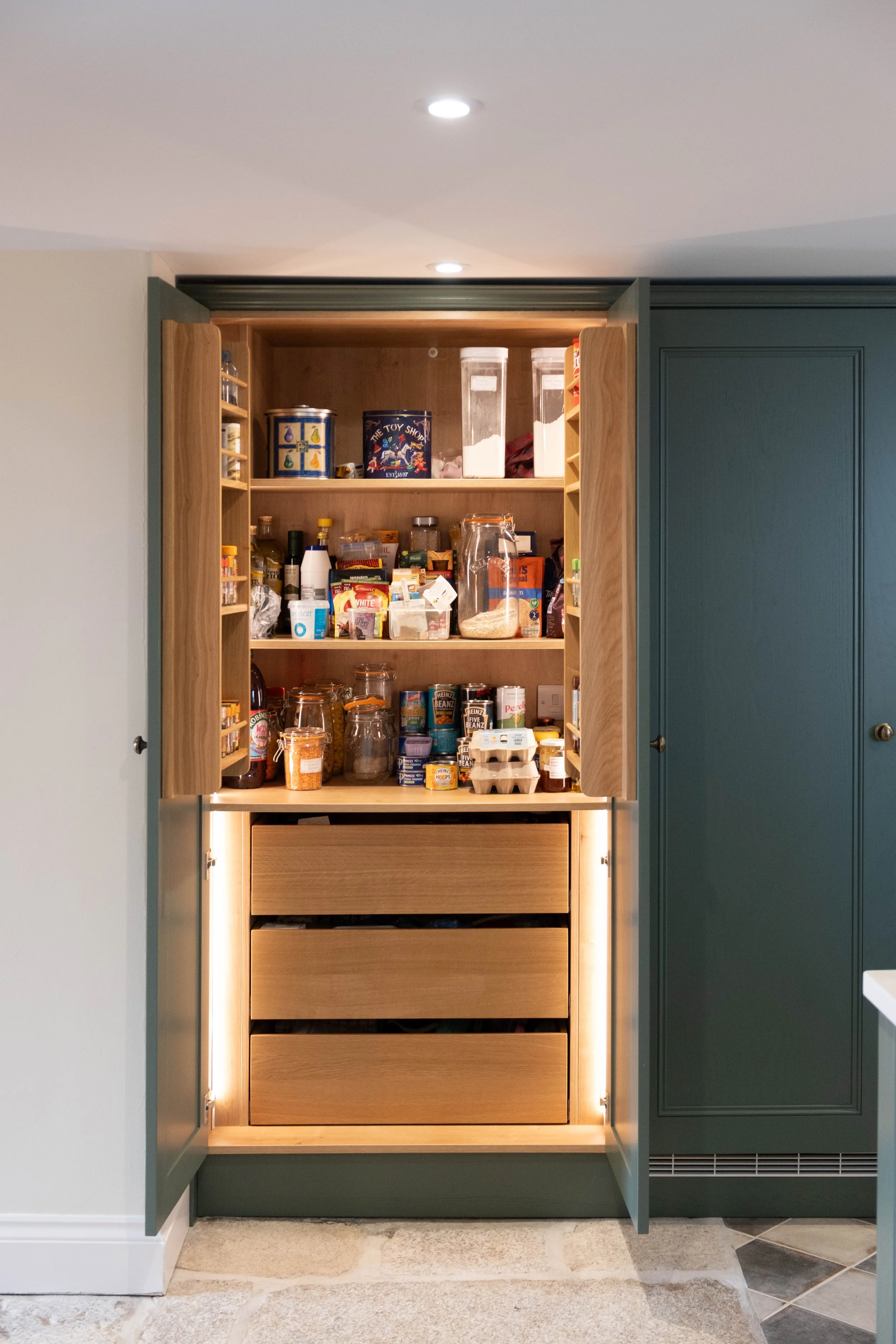 Open kitchen cabinet with shelves filled with various food items and jars, and lower drawers.