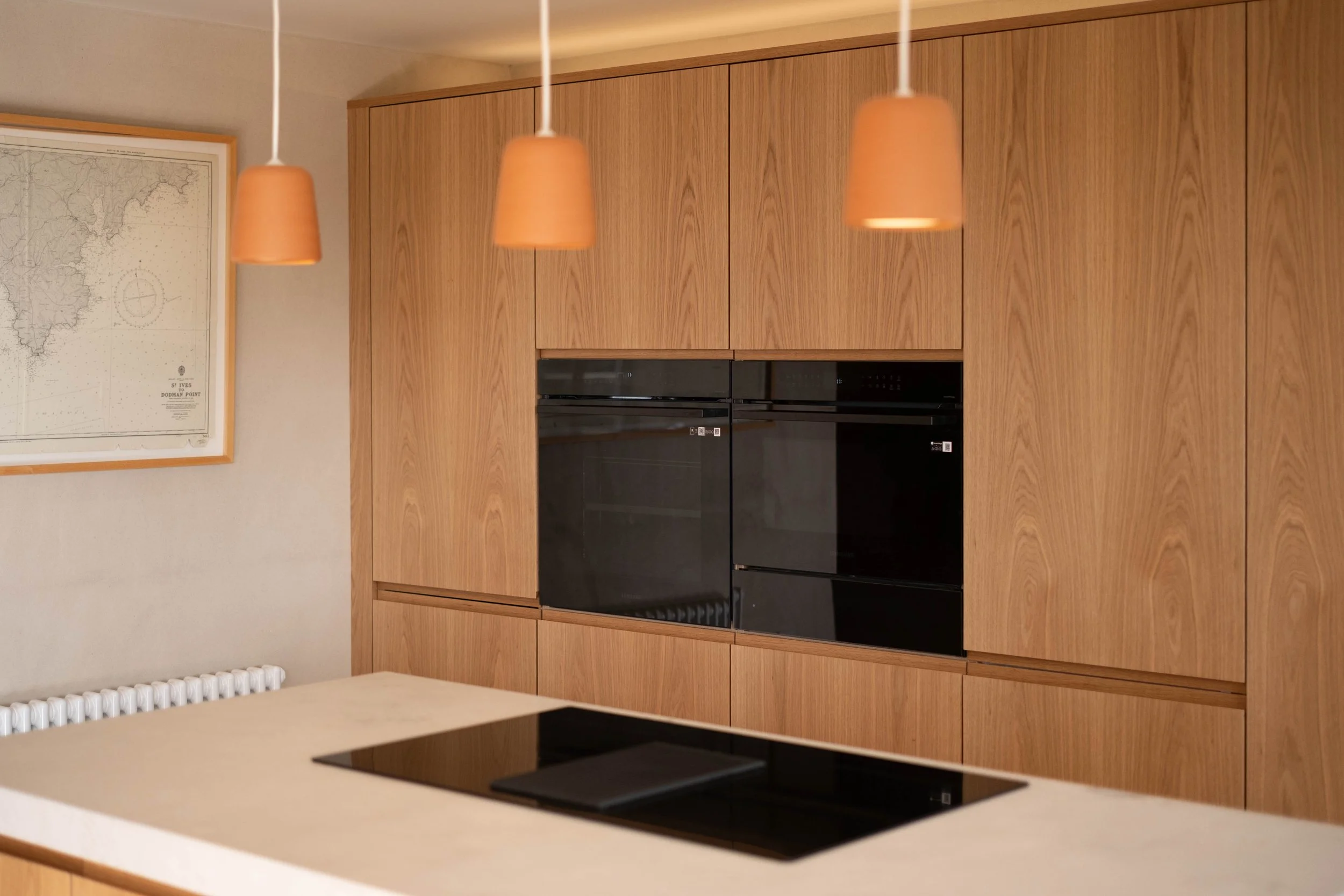 Modern kitchen with wooden cabinetry, black oven, and beige countertops, illuminated by three pendant lights.