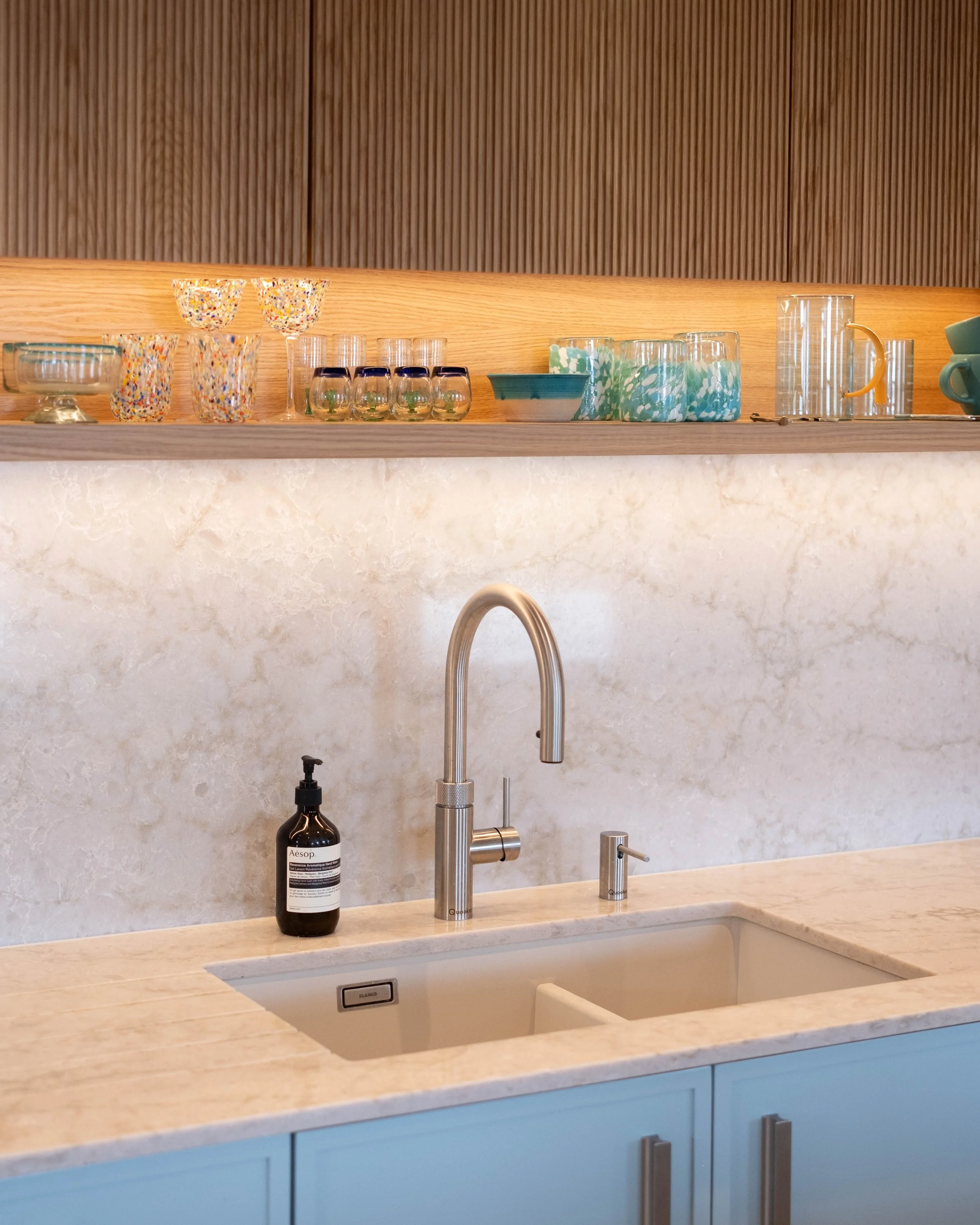 A kitchen countertop with a sink, a black pump bottle of Aesop hand soap, a modern chrome faucet, glasses, bowls, and cups, and blue cabinets underneath, with a textured backsplash and wooden wall above.
