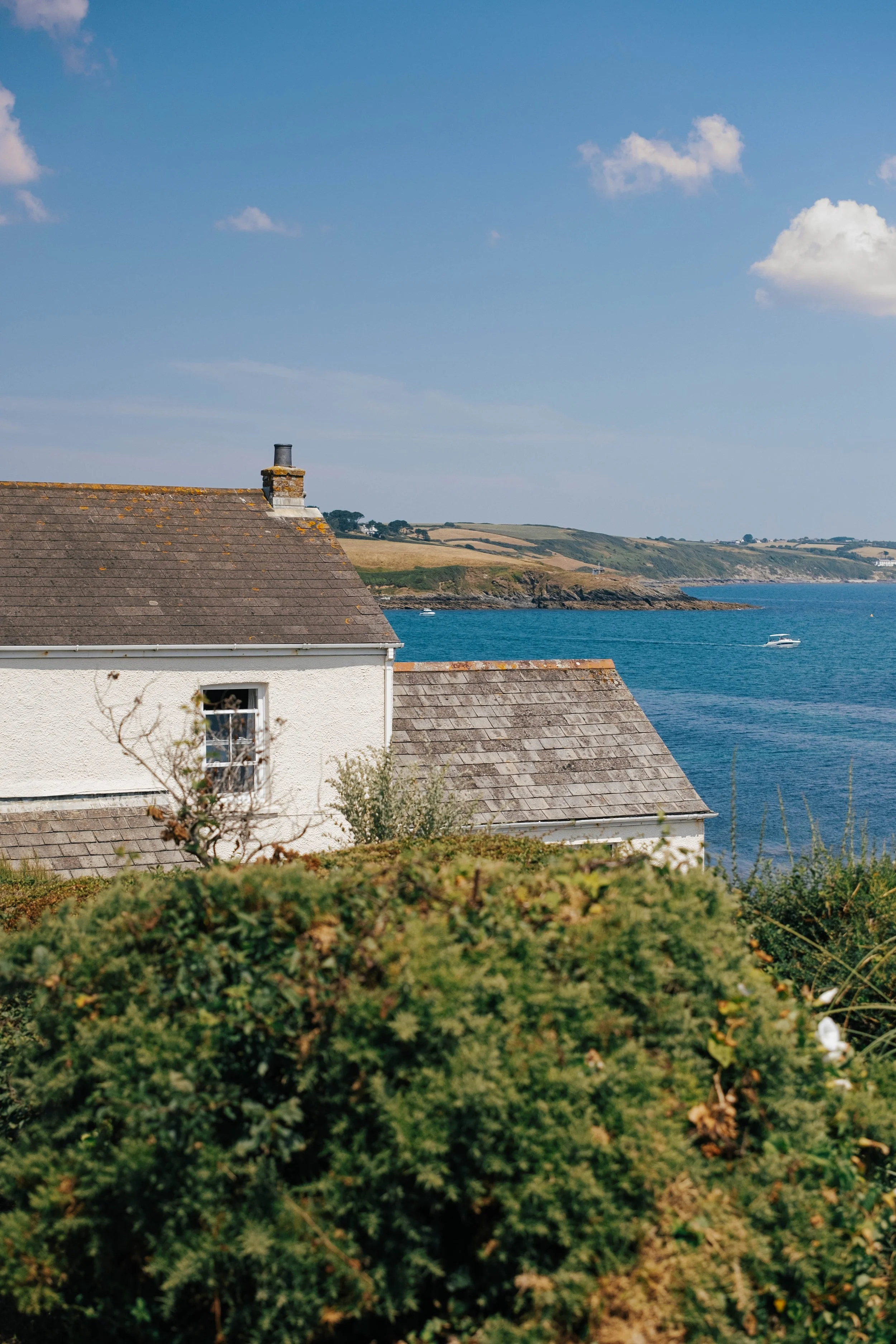 A house with a tiled roof situated near the coastline, with the sea and hills in the background under a partly cloudy sky.