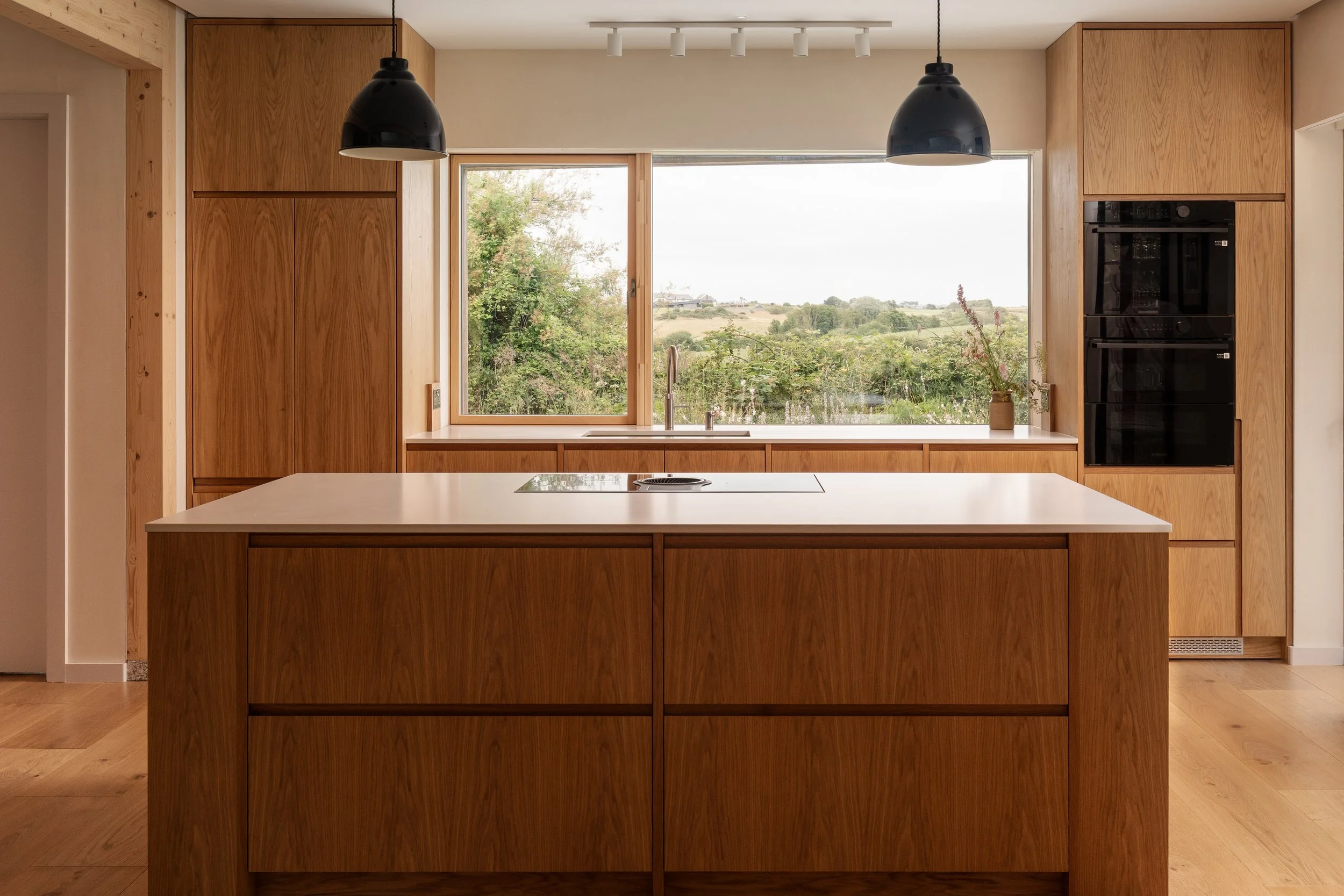 Modern kitchen with wooden cabinets, a large window showcasing a scenic landscape, and two black pendant lights hanging above a kitchen island.