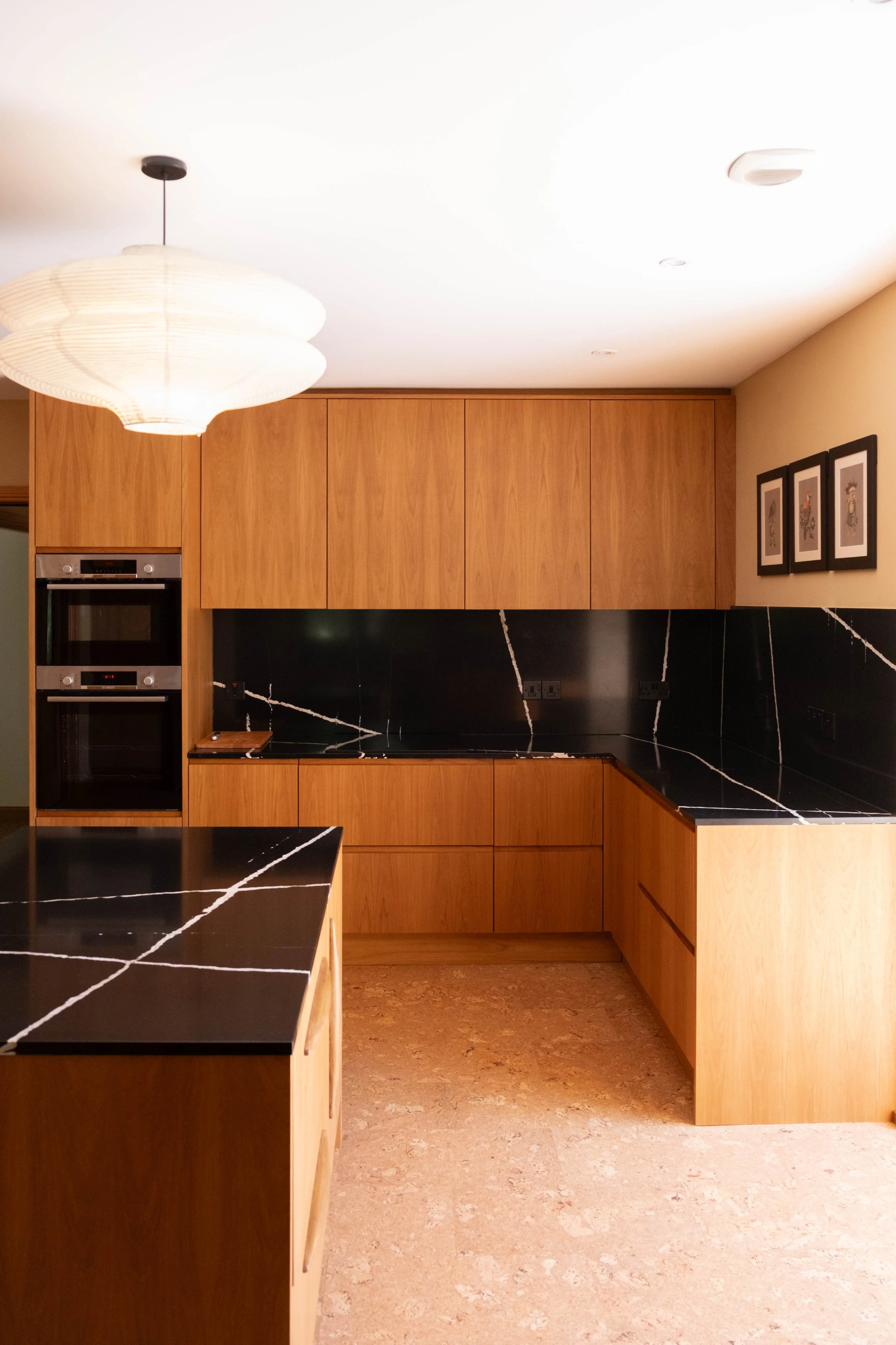 A modern kitchen with wooden cabinets, black marble countertops with white veining, a built-in oven, and a hanging paper lantern light fixture.
