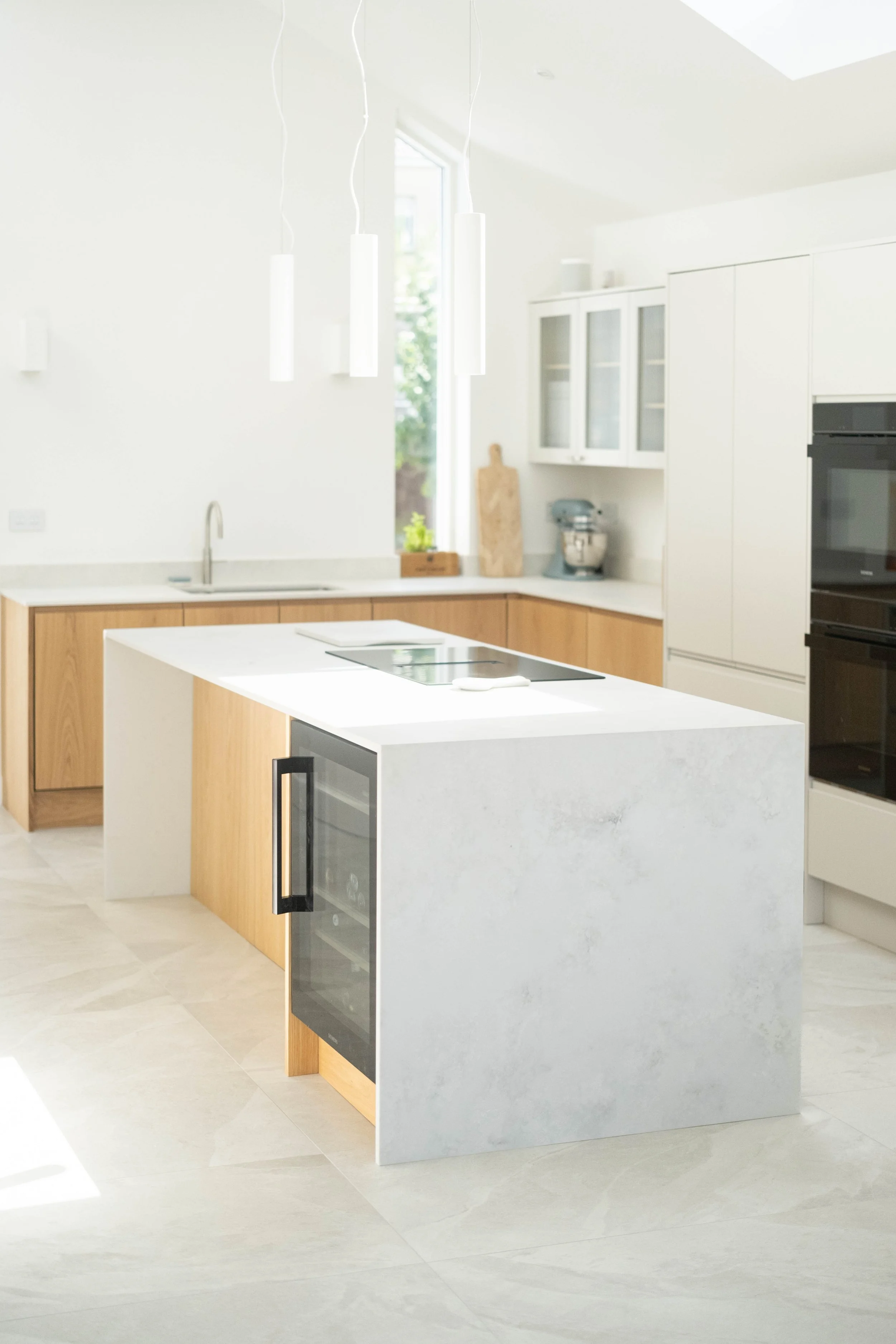 Modern kitchen with white countertops, light wooden cabinets, a built-in wine cooler, a black oven, a sink, hanging pendant lights, and large windows letting in natural light.