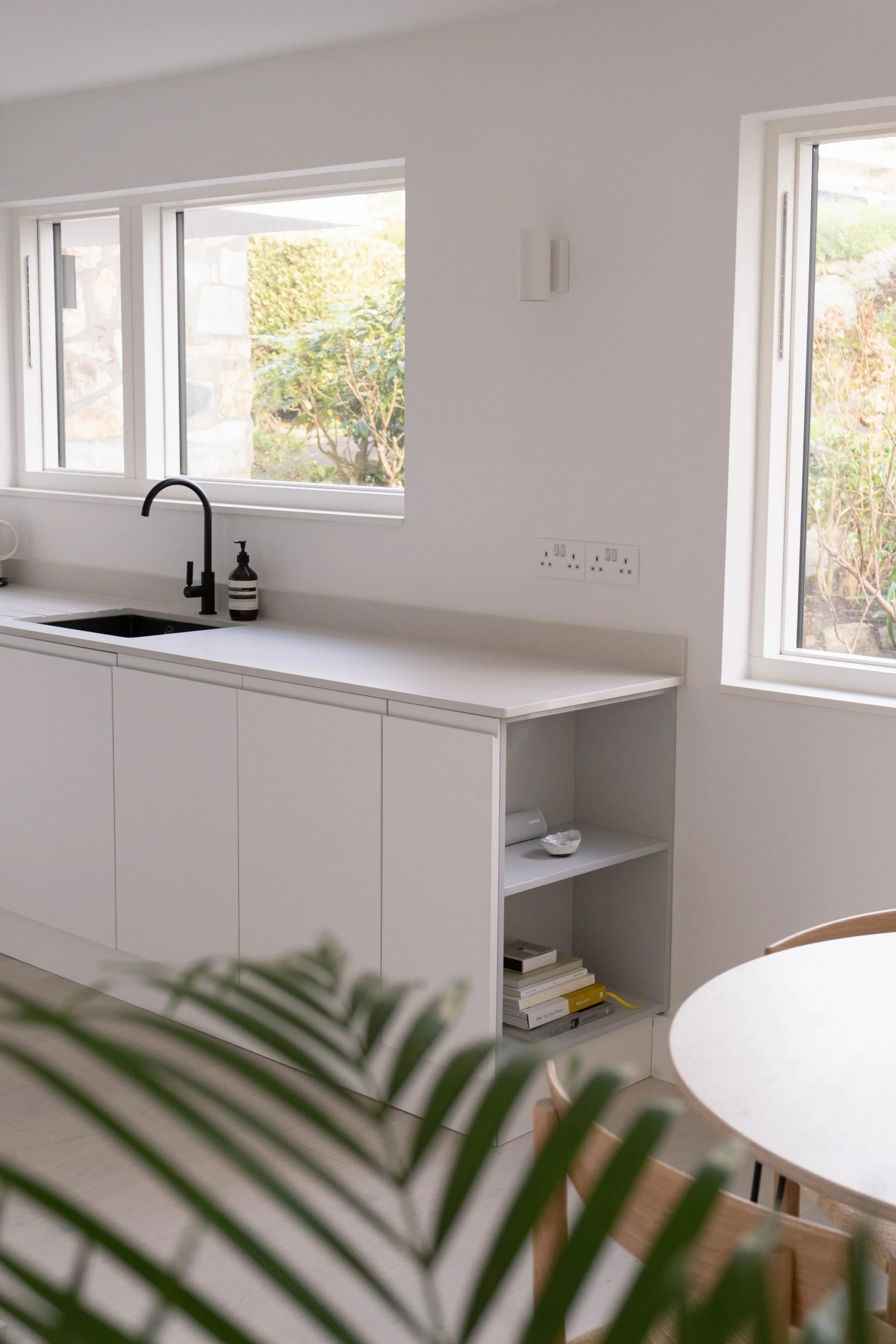 Interior of a minimalist kitchen with white cabinets, black faucet, window overlooking greenery, small open shelves with books, and a round dining table with wooden chairs.