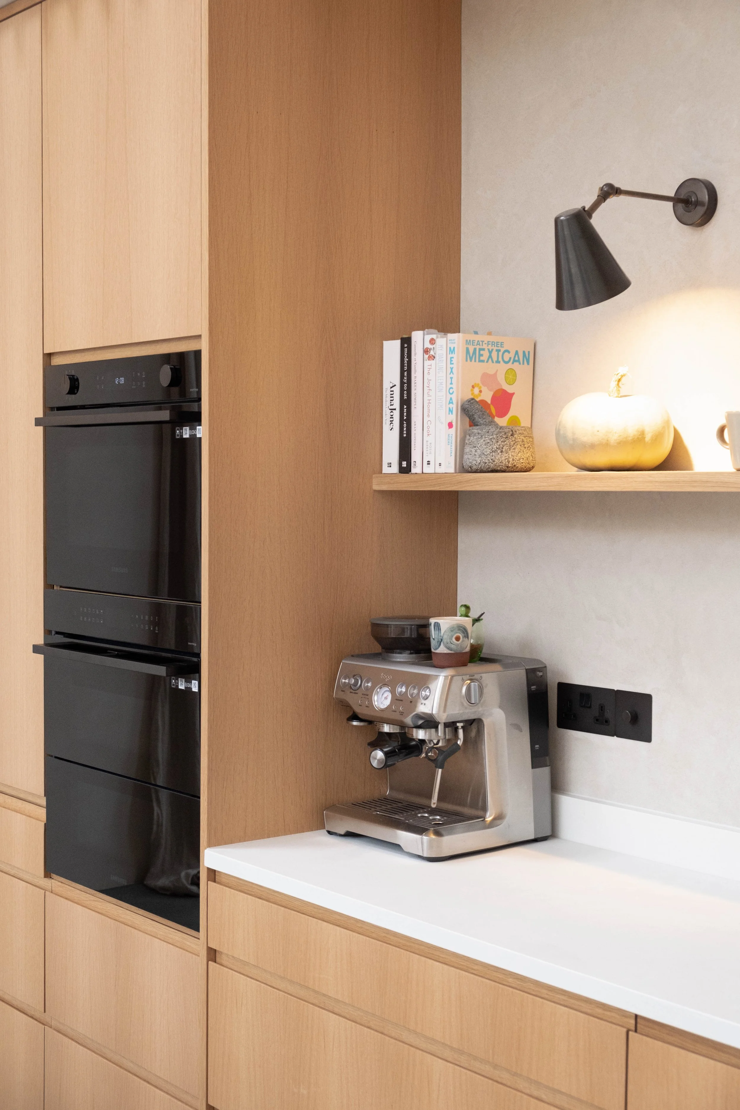 A modern kitchen with built-in oven, wooden cabinets, a white countertop, a coffee machine, a small shelf with books, a decorative pumpkin, a mortar and pestle, and a wall-mounted lamp.