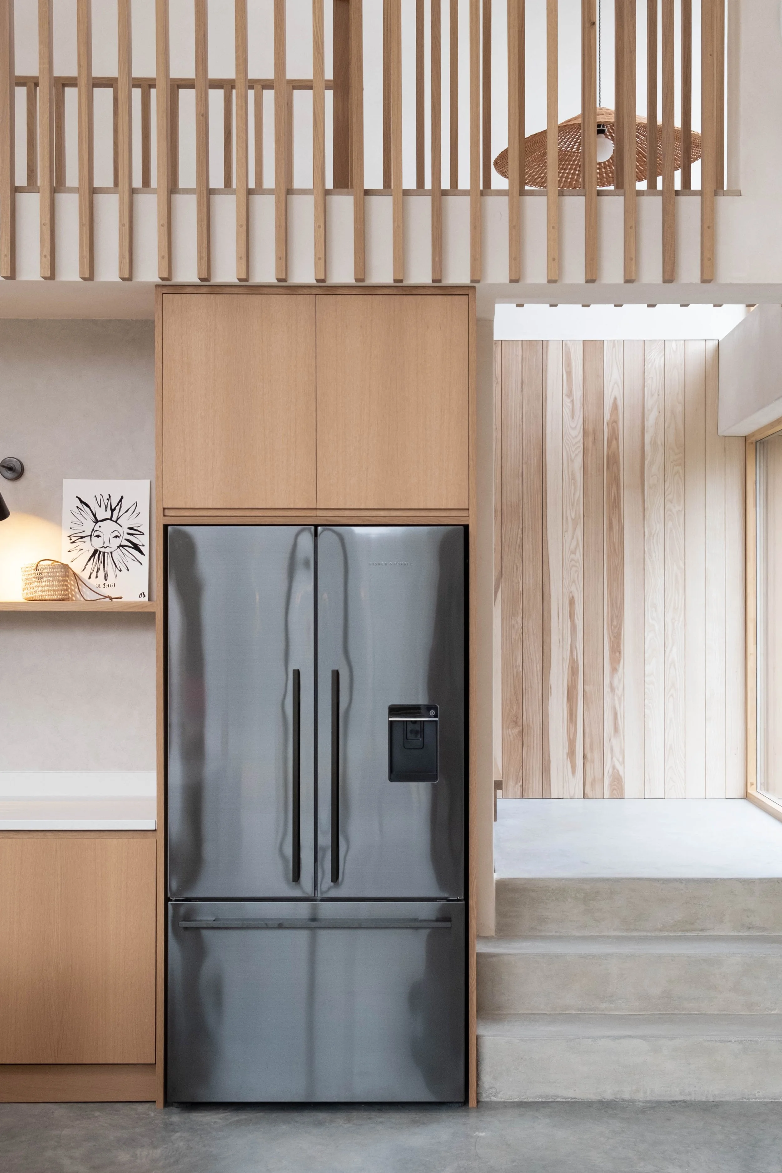 Interior view of a modern kitchen with wooden cabinetry, stainless steel refrigerator, and light wood wall paneling. A staircase with concrete steps is visible to the right.