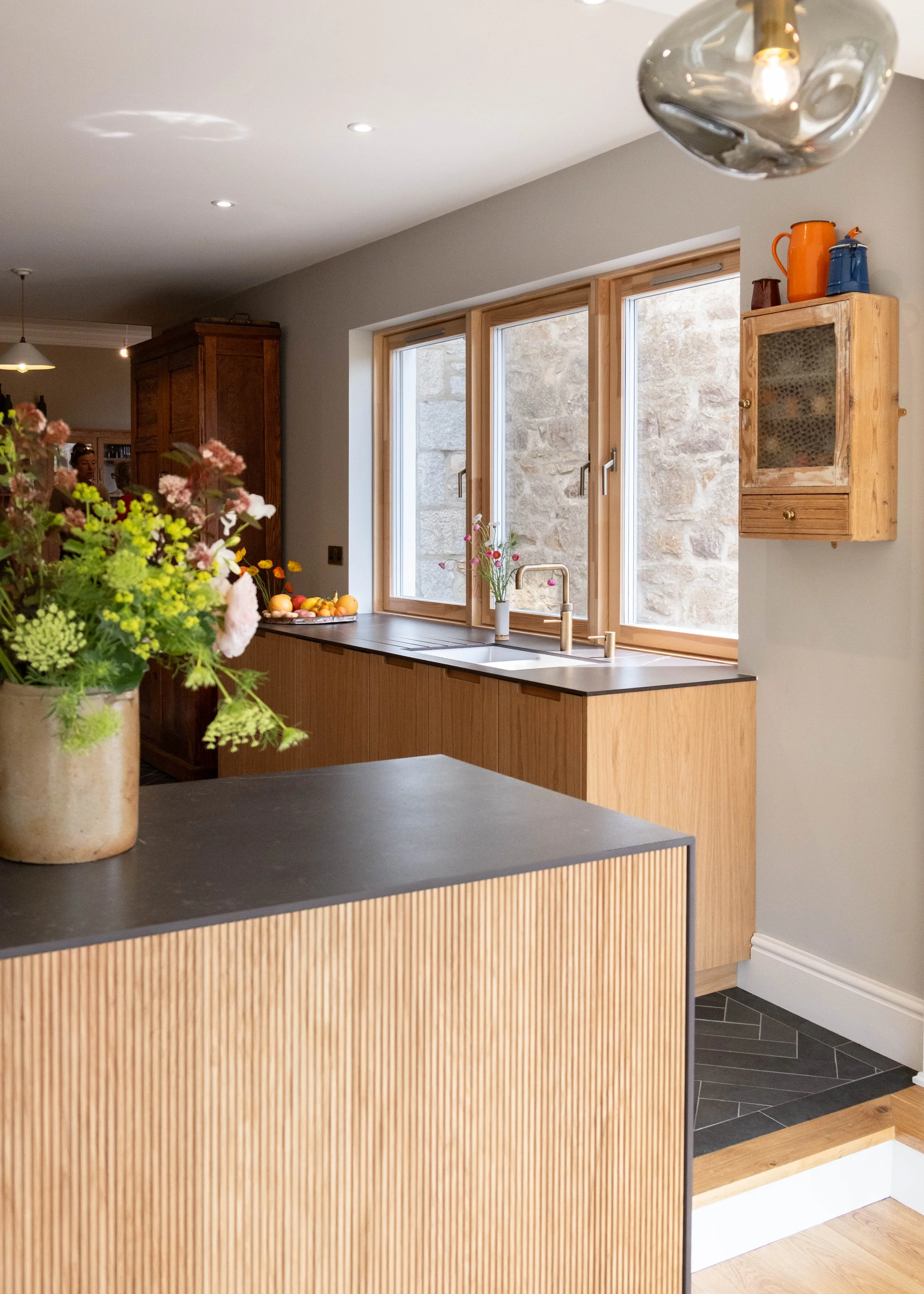 Kitchen with a black island counter, a vase of flowers, a window with a stone wall outside, a wooden wall cabinet with colorful mugs, and a textured wooden panel on the counter front.