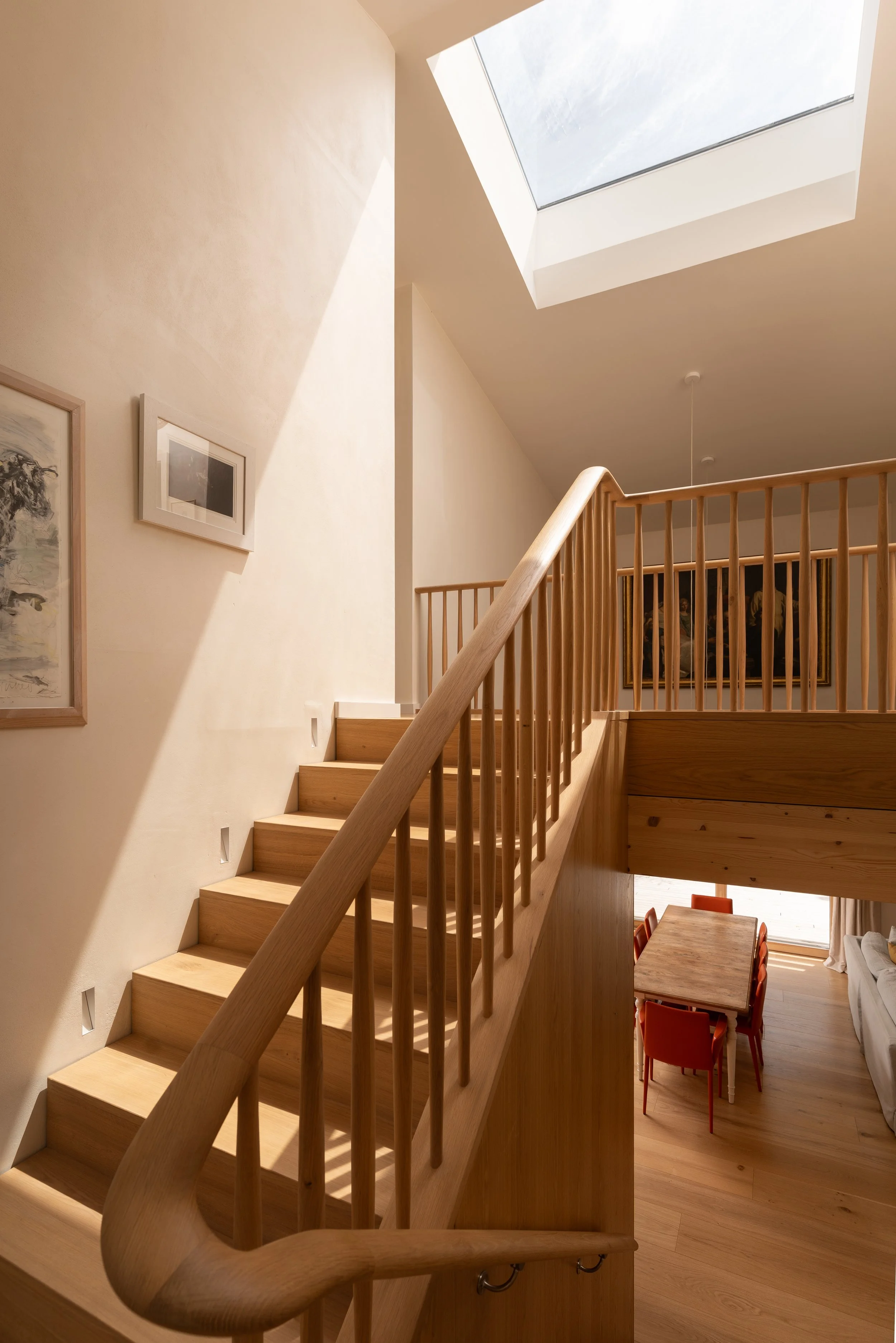 Wooden staircase with curved handrail leading to a lower dining area, skylight ceiling with natural light, beige walls with framed artwork, and a glimpse of a bench or sofa with cushions.