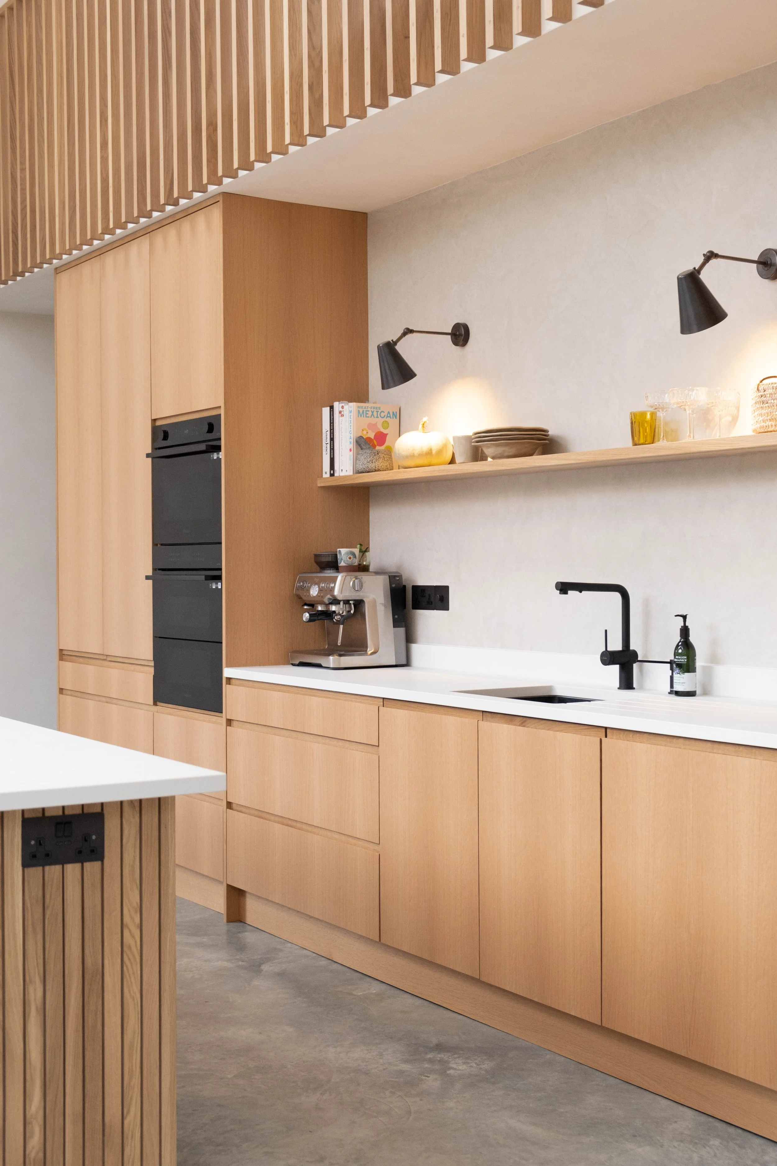 Modern kitchen with light wood cabinets, black wall-mounted lamps, a white countertop, and a built-in black oven. Decor includes a pumpkin, books, plates, glasses, and a coffee machine.