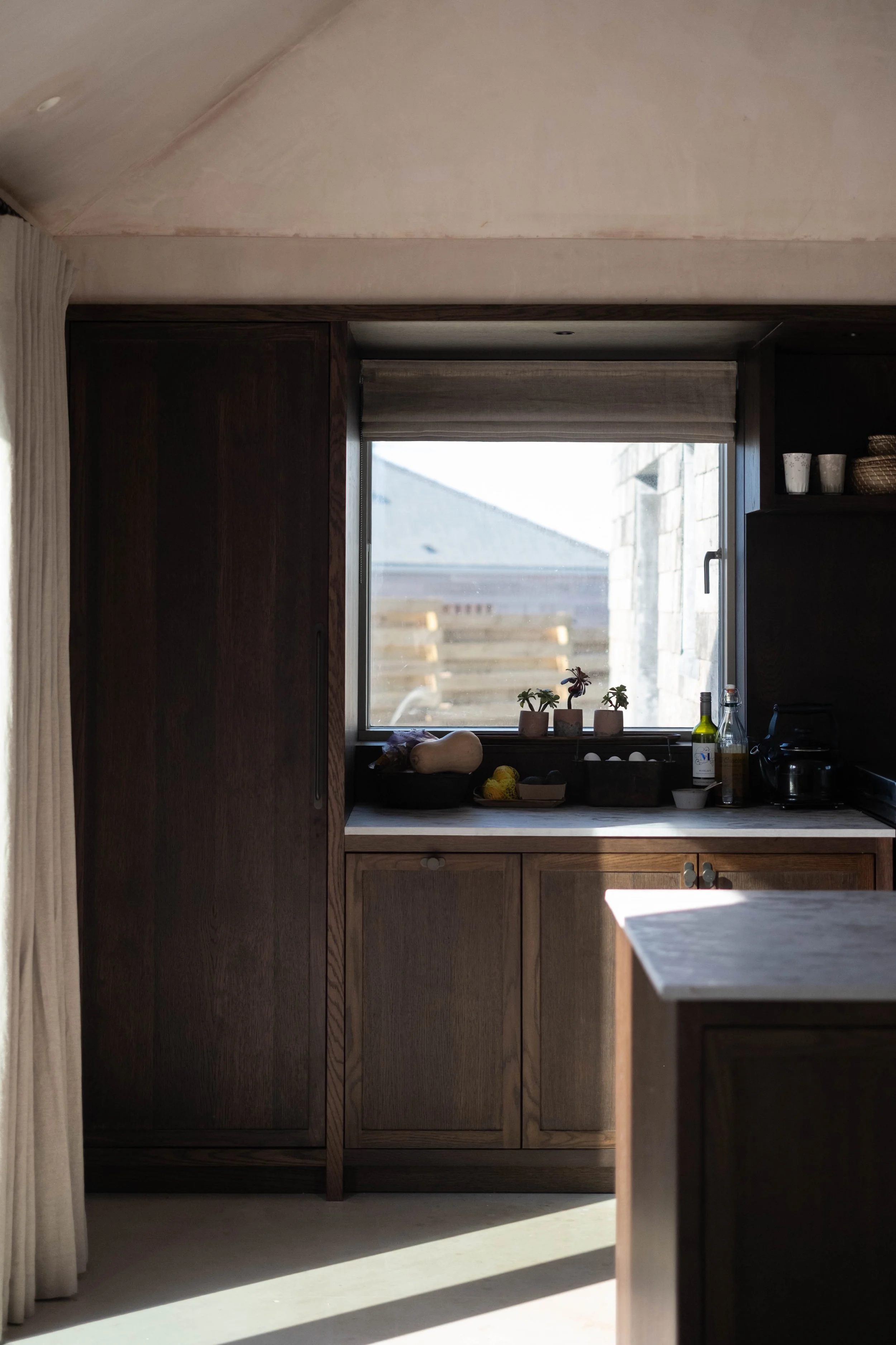 A kitchen with wooden cabinets, a window with a view of the outdoors, and various kitchen items such as potted plants, bottles, and containers. Sunlight is coming through the window.