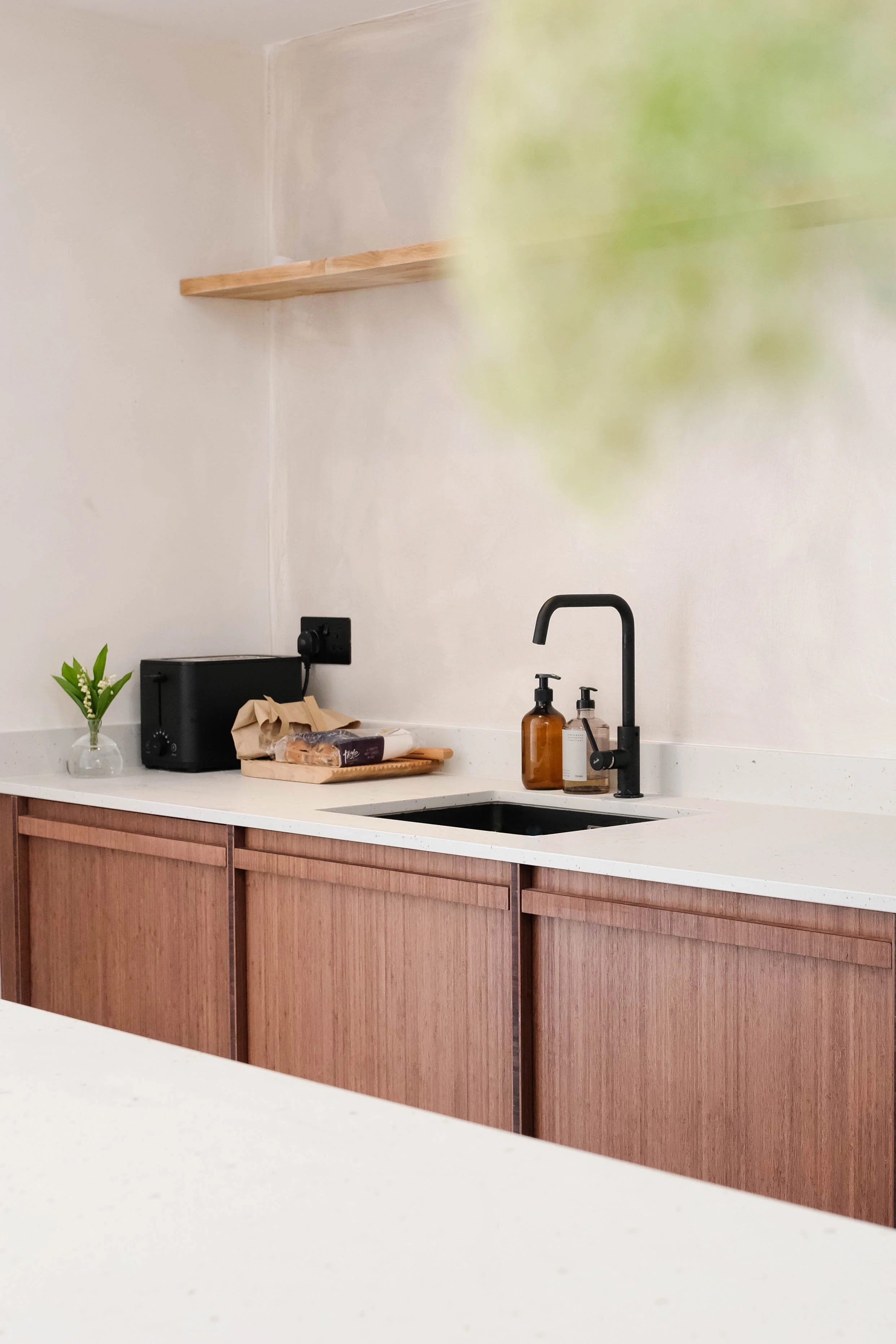 Kitchen countertop with a small potted plant, a black toaster oven, a paper bag, and some food items. The background shows a sink area with brown bottles and a black faucet, with a wooden cabinet below and a plain white wall with a floating wooden shelf.