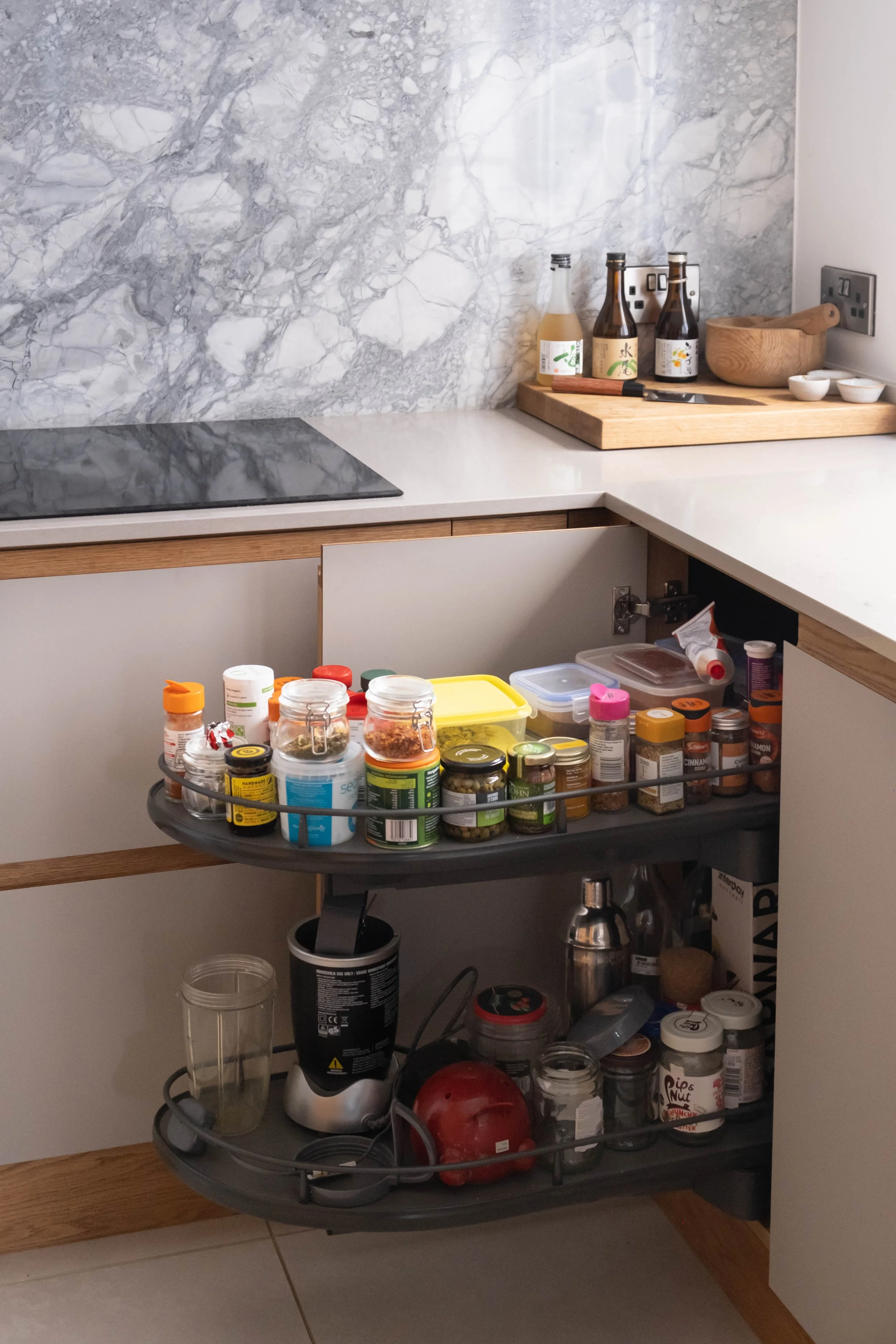 Kitchen countertop with bottles of seasoned sauce, a wooden bowl with a spoon, small bowls, and a cutting board near a wall outlet. Below, a cart holds jars of spices and supplements, a blender, and various kitchen items.