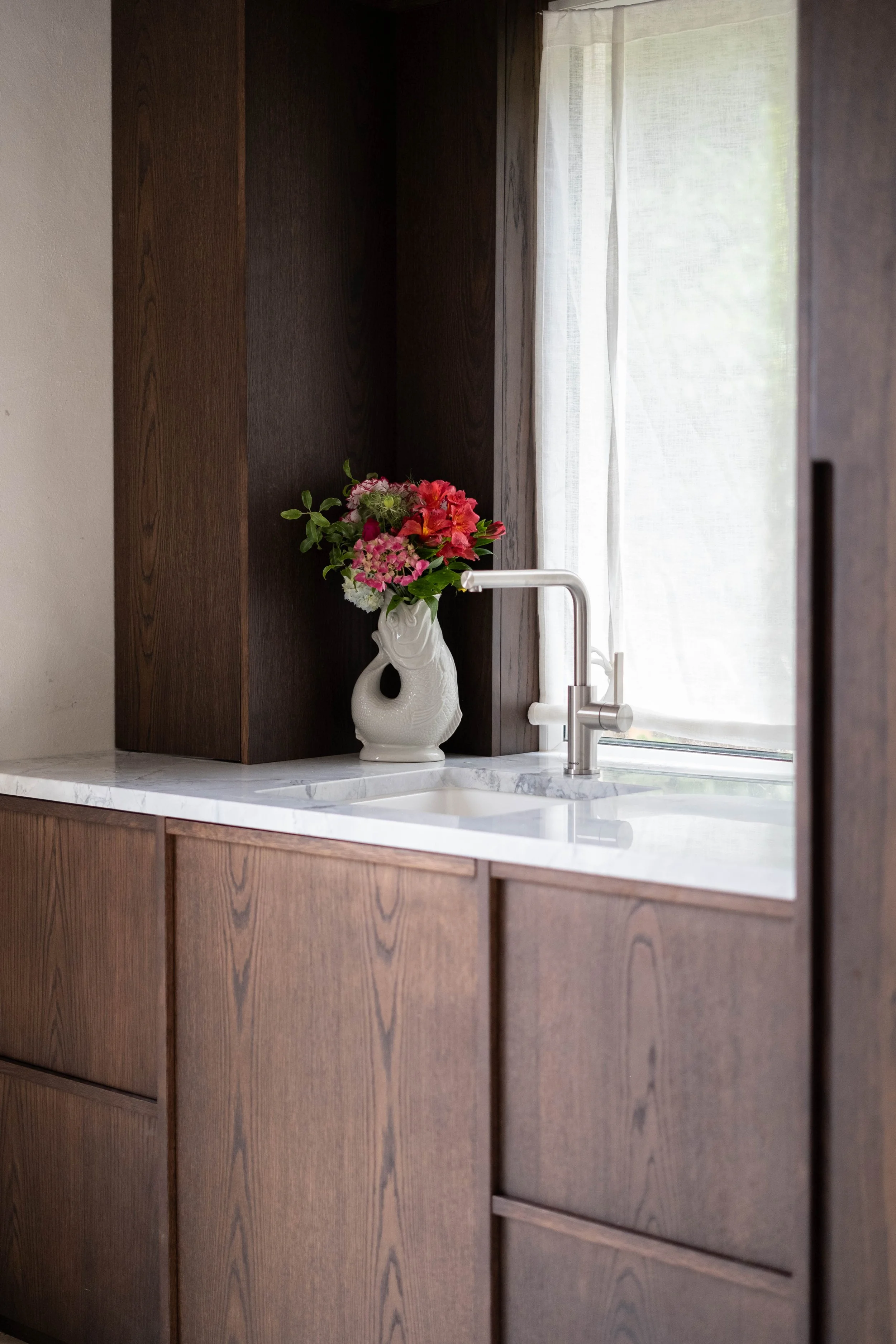 A kitchen window with white curtains, a white ceramic vase with pink and red flowers, a stainless steel faucet, and brown wooden cabinetry with a marble countertop.