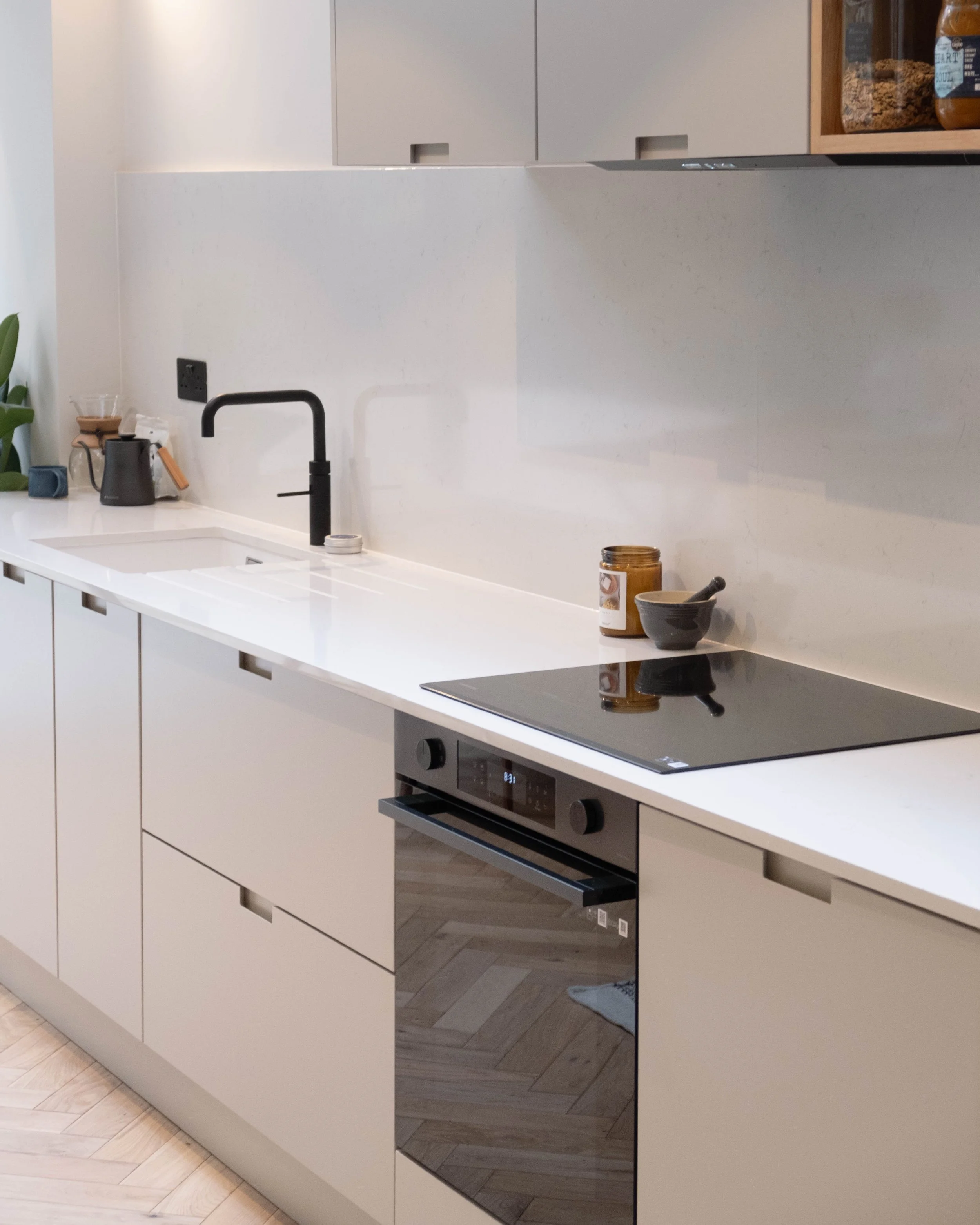 Modern white kitchen with a black faucet, a black kettle, a potted plant, and various jars on the countertop. There is a built-in oven and a sleek black induction cooktop, with light wood flooring and minimalistic cabinetry.