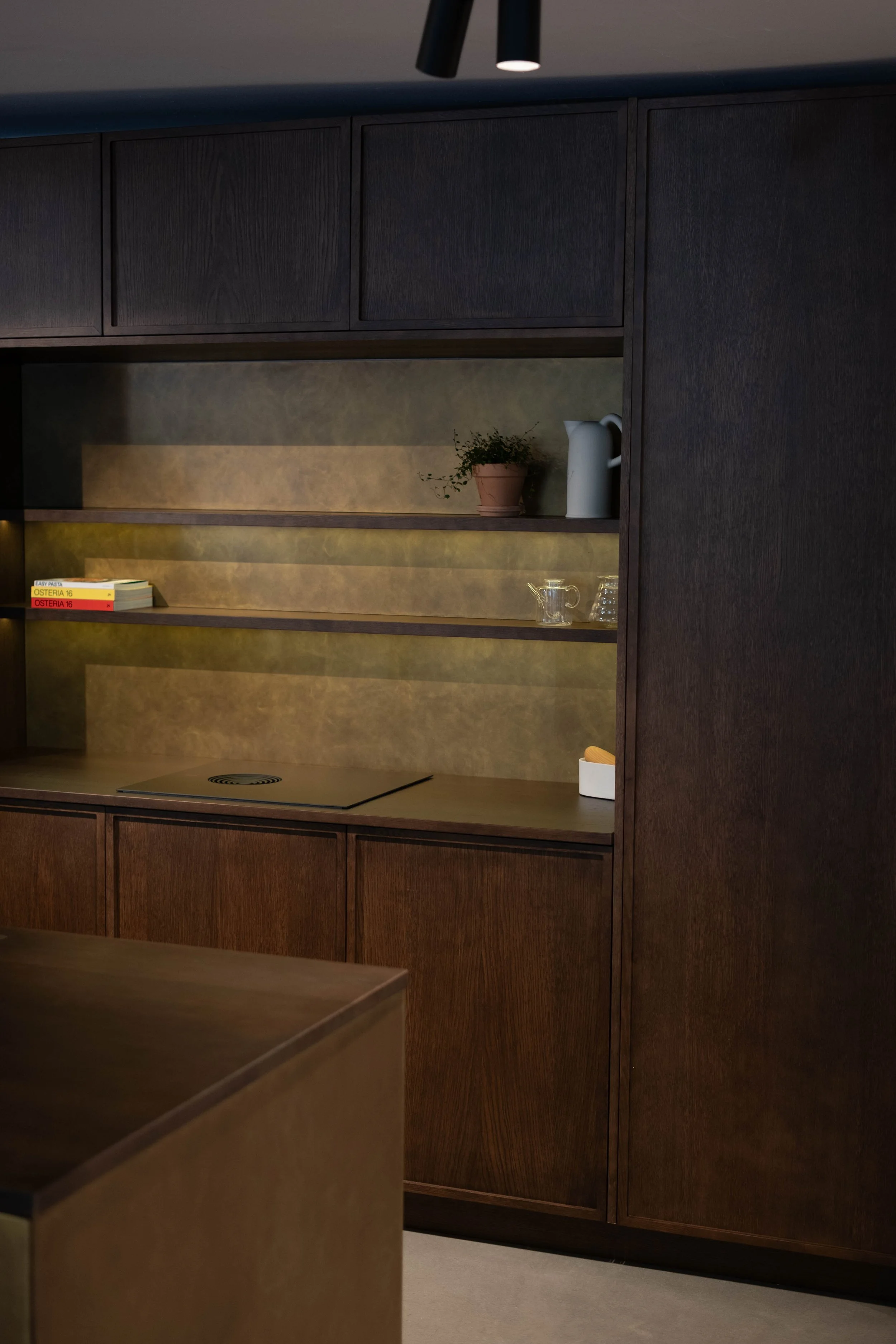 A kitchen with dark wood cabinets and a built-in shelf that has a potted plant, a white pitcher, and glassware, with a stovetop visible on the counter.