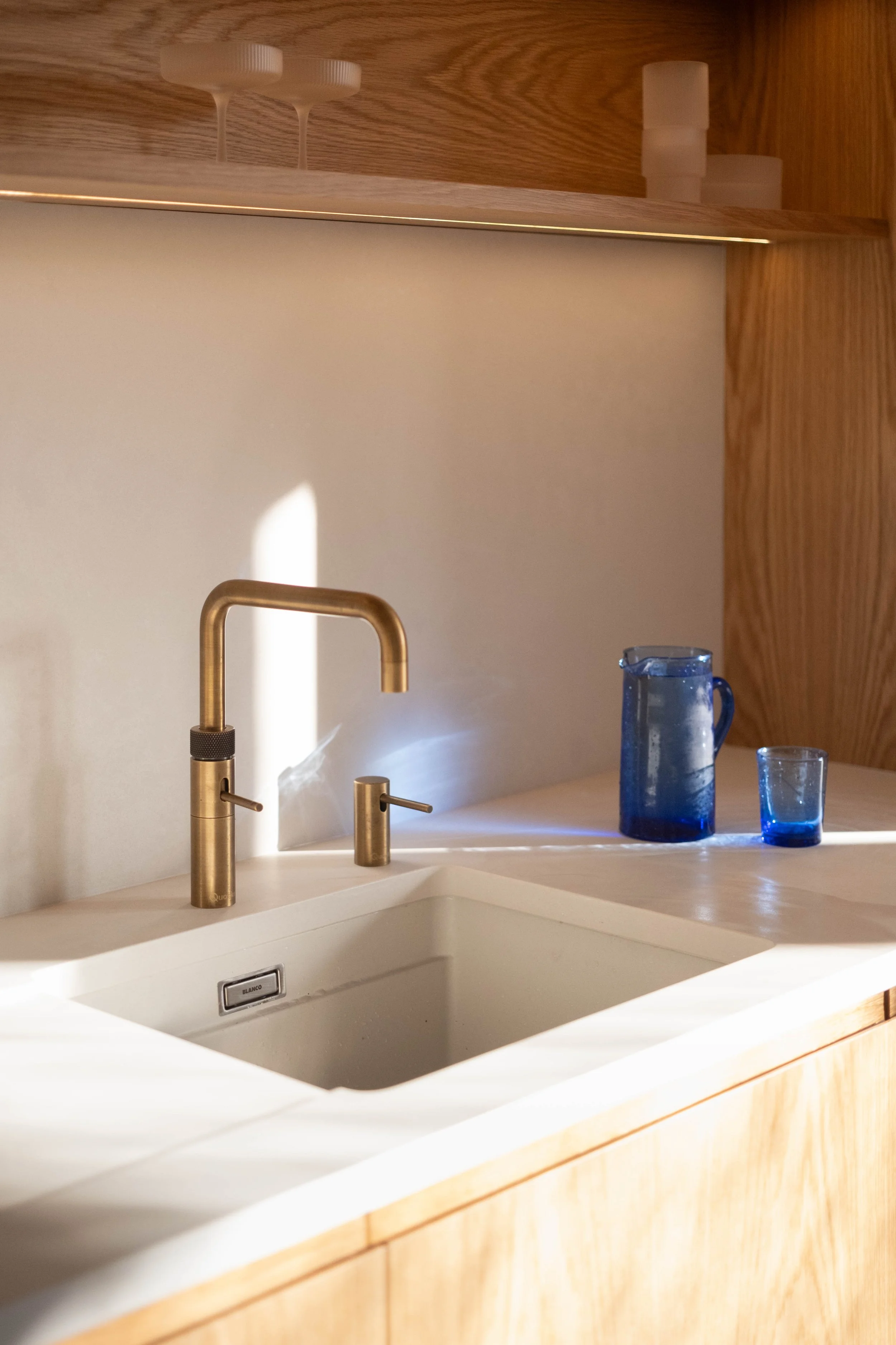 A modern kitchen sink with a gold faucet, a small soap dispenser, and two blue glass pitchers on the counter, with wooden cabinets and a white wall in the background.