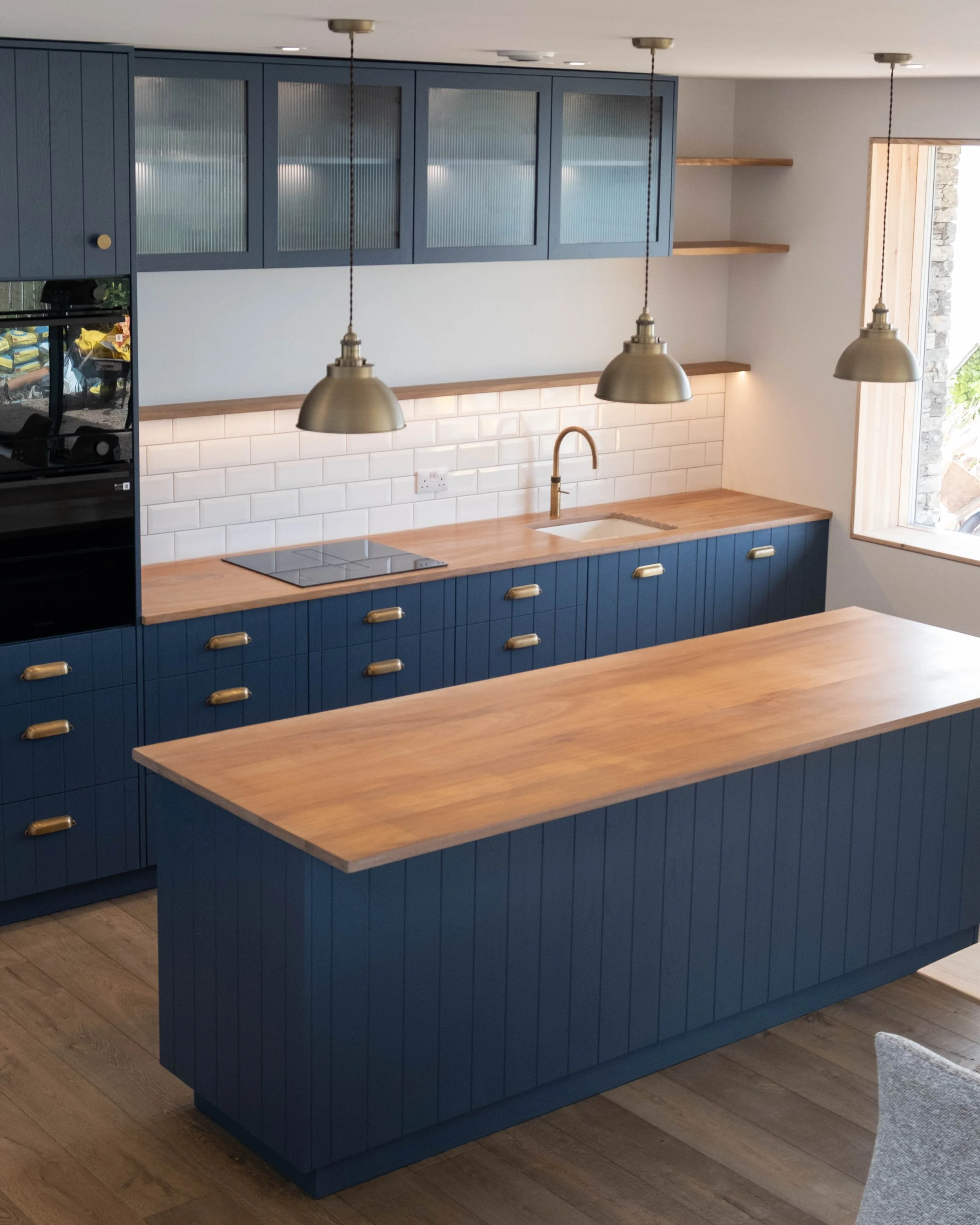 A modern kitchen with blue cabinets, wooden countertops, white subway tile backsplash, and a window with natural light. There are three pendant lights hanging above the counter.