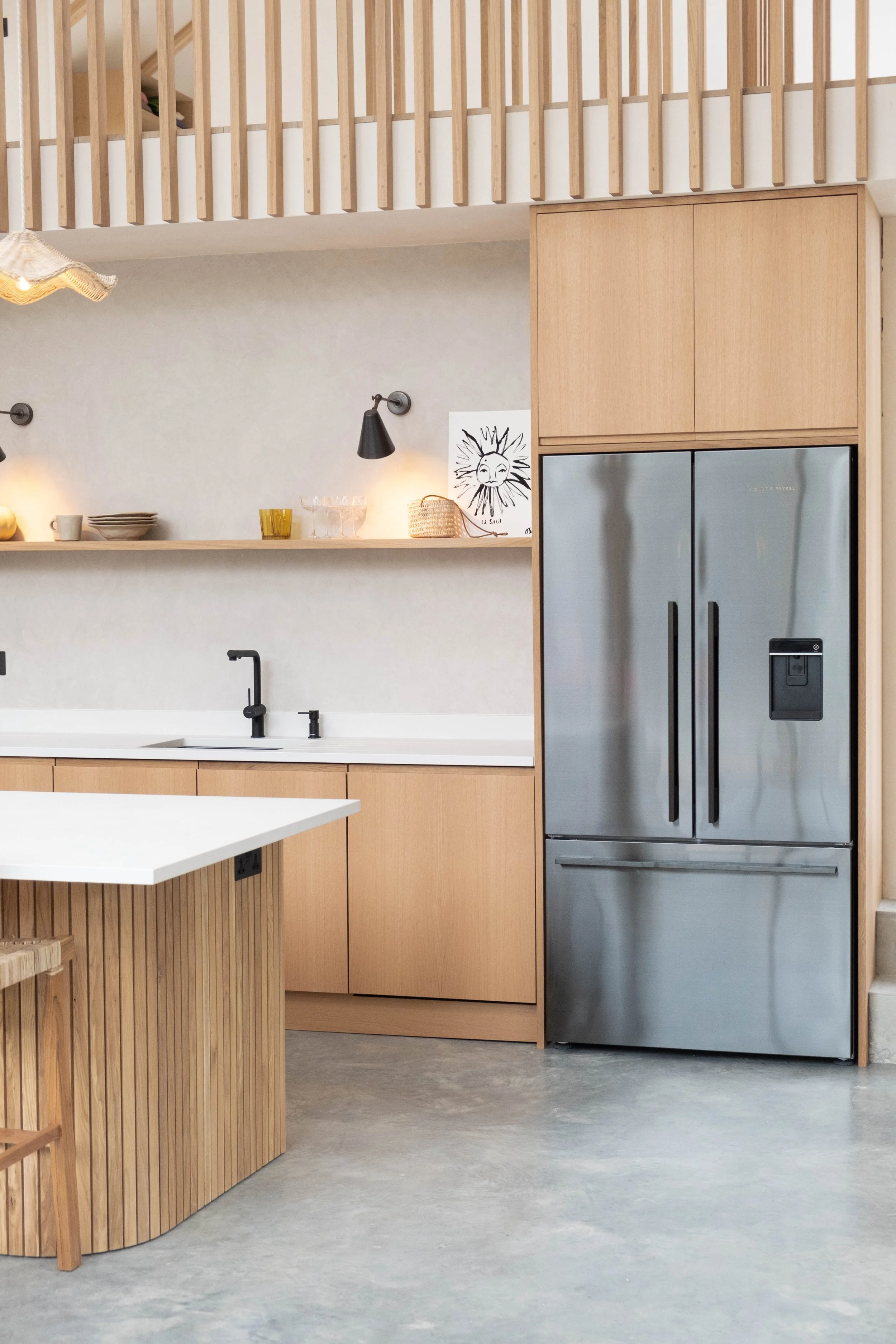 Modern kitchen with light wood cabinets, stainless steel refrigerator, black faucet, open shelves with cups and bowls, wall decor of a sun with a face, and a wooden island with vertical slats.