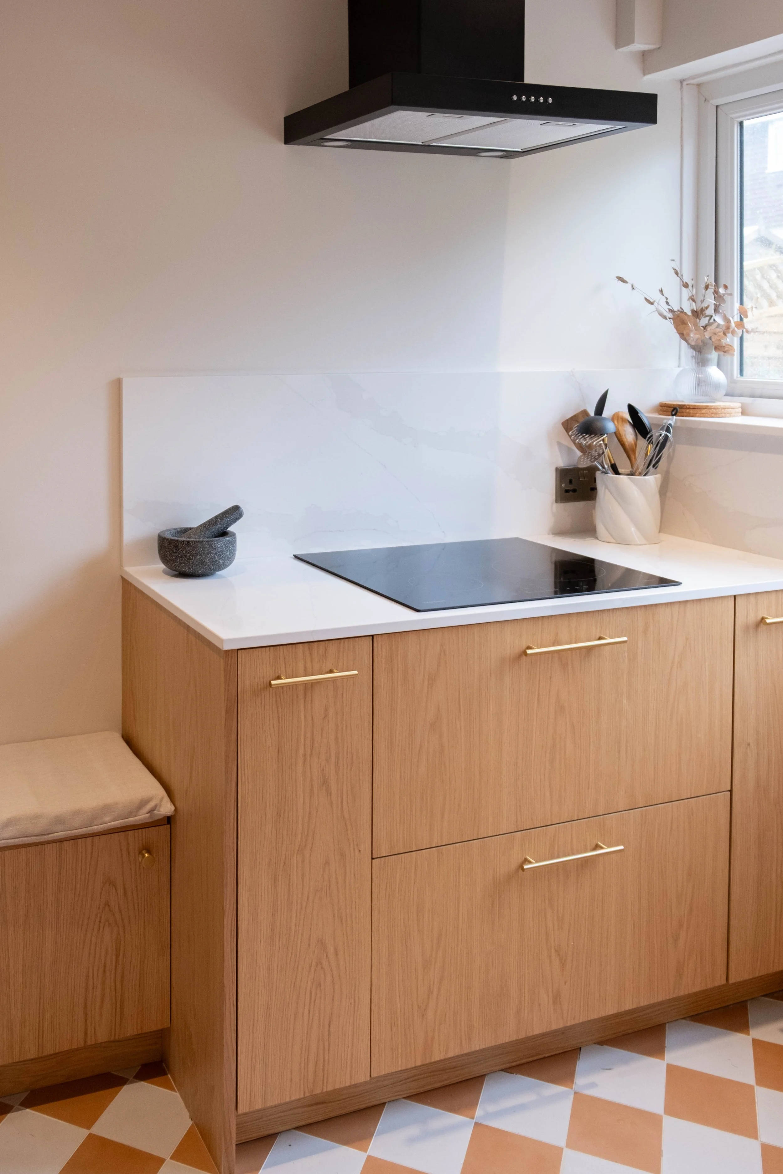 A modern kitchen countertop with a black electric cooktop, a mortar and pestle, and a container holding kitchen utensils. There is a window with a vase of dried flowers on the windowsill and a beige cushioned seat with gold handles in the corner. The