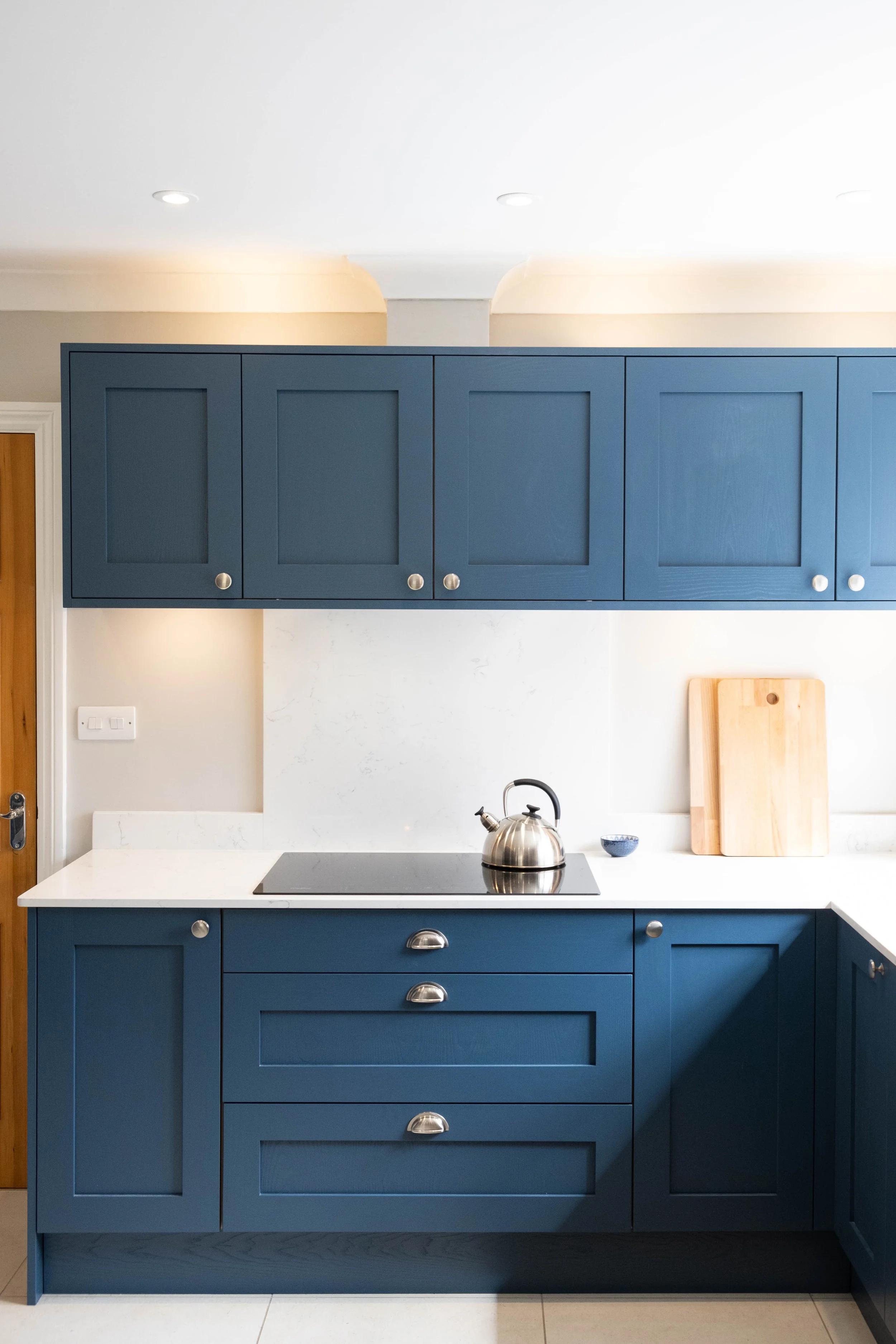 Blue kitchen cabinets above and below a white countertop with a stovetop. A silver kettle and a small bowl are on the counter. Two wooden cutting boards lean against the wall. Recessed lights are on the ceiling.