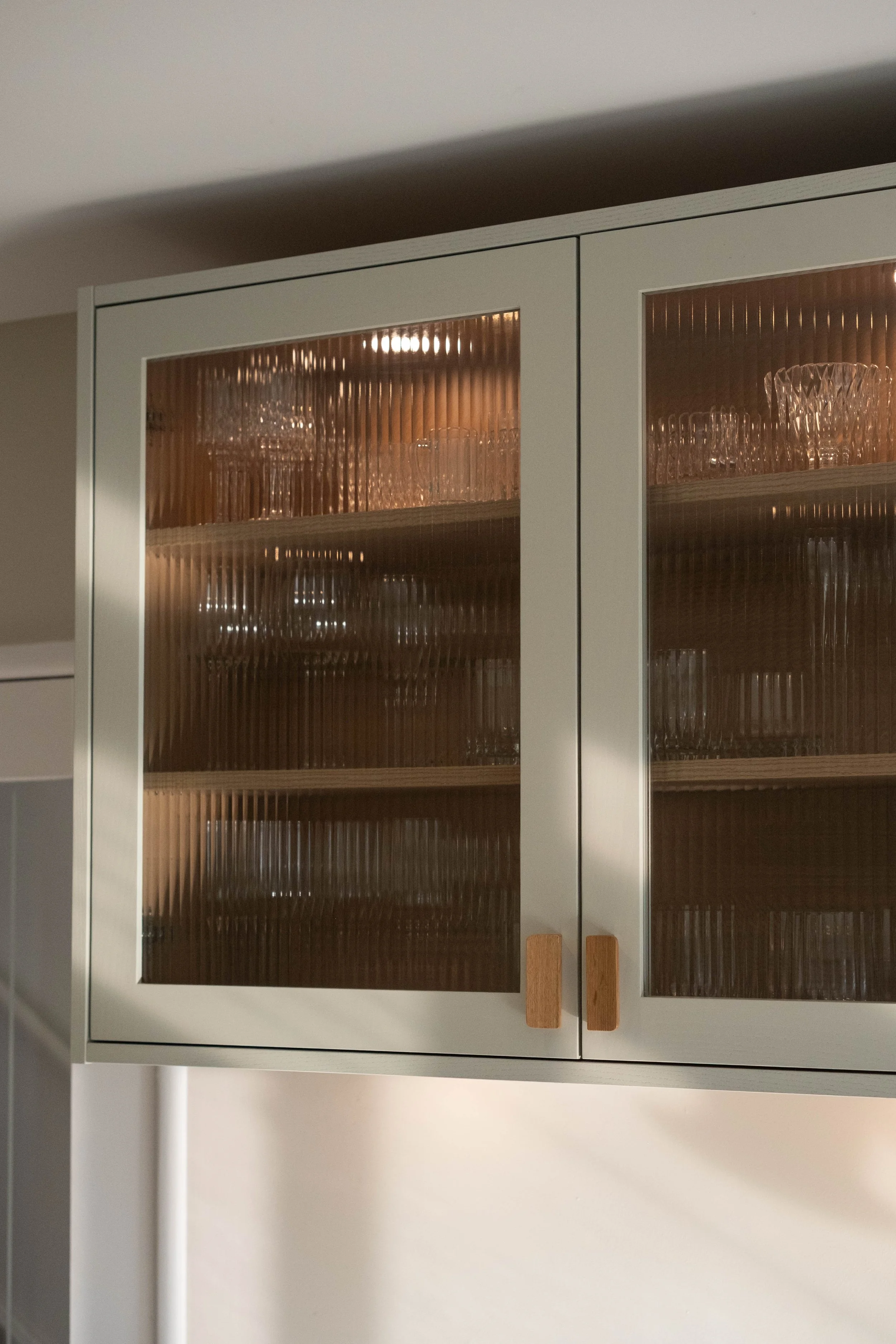 A white kitchen cabinet with glass doors, containing glassware inside, and two wooden handles. The cabinet is against a beige wall, with a white ceiling above.