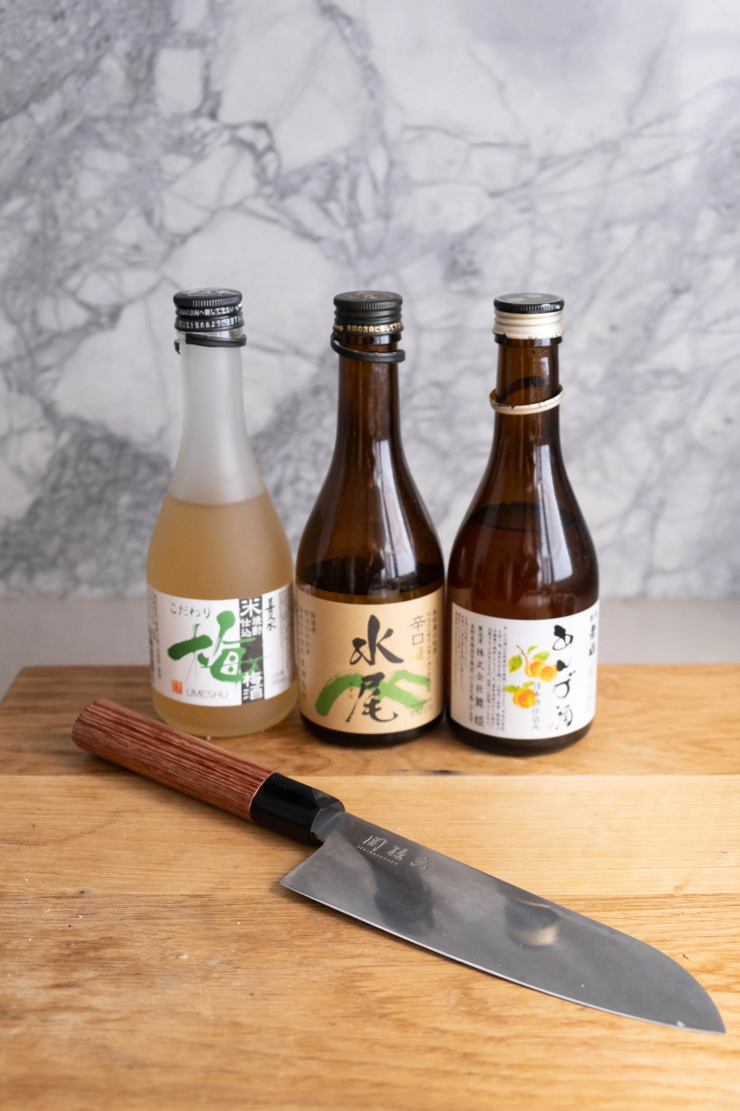 Three bottles of Japanese beverages on a wooden surface with a knife in front, against a marble wall background. The bottles are different colors and labels with Japanese writing.