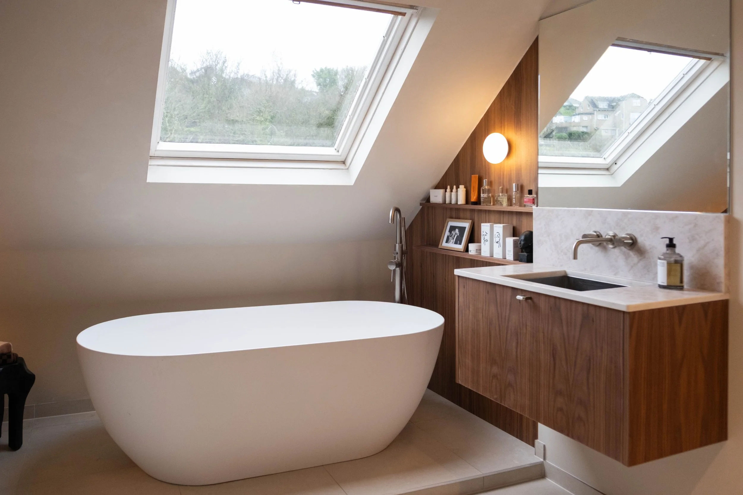 Modern bathroom with skylight windows, a freestanding bathtub, wooden cabinetry, and various toiletries on shelves visible in the mirror reflection.