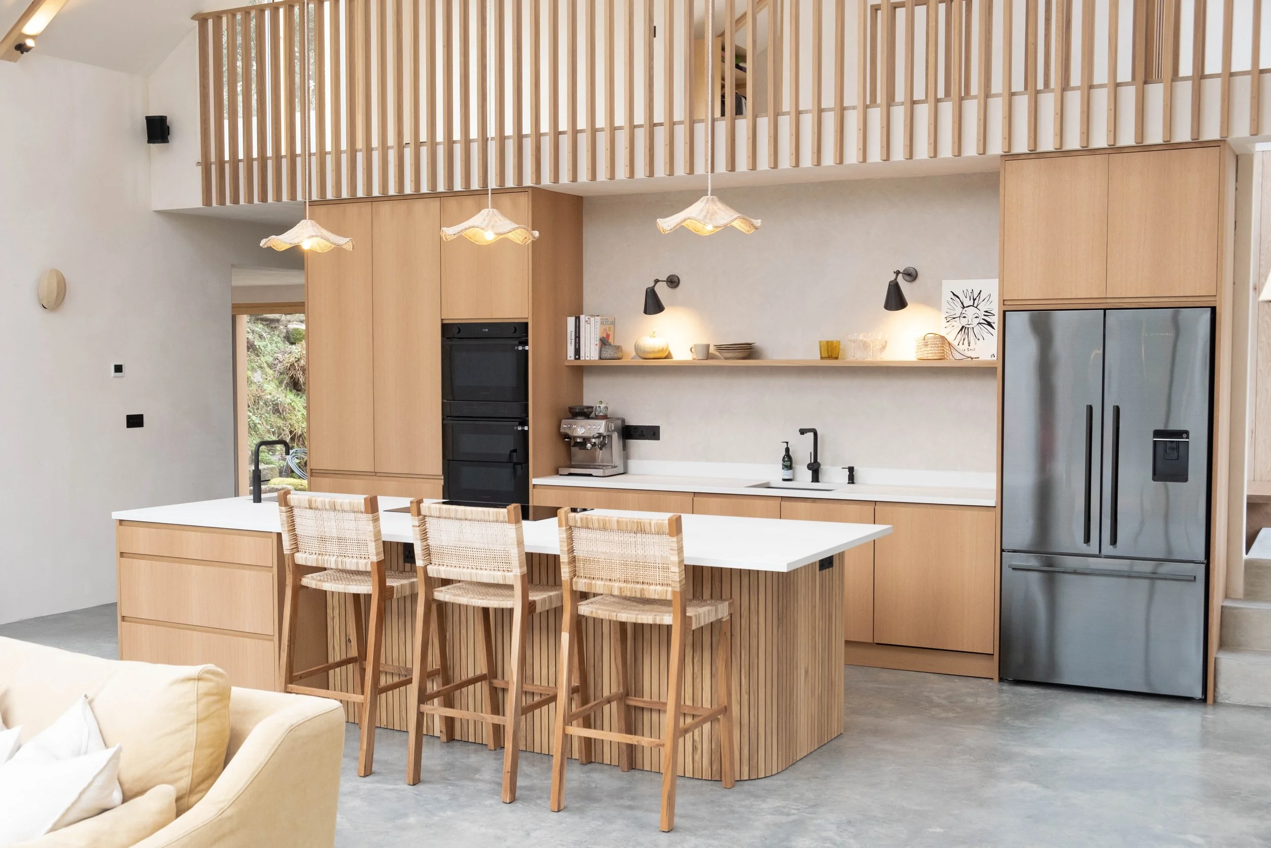 Modern kitchen with light wood cabinets, a white island with chairs, pendant lights, and a view of greenery outside.