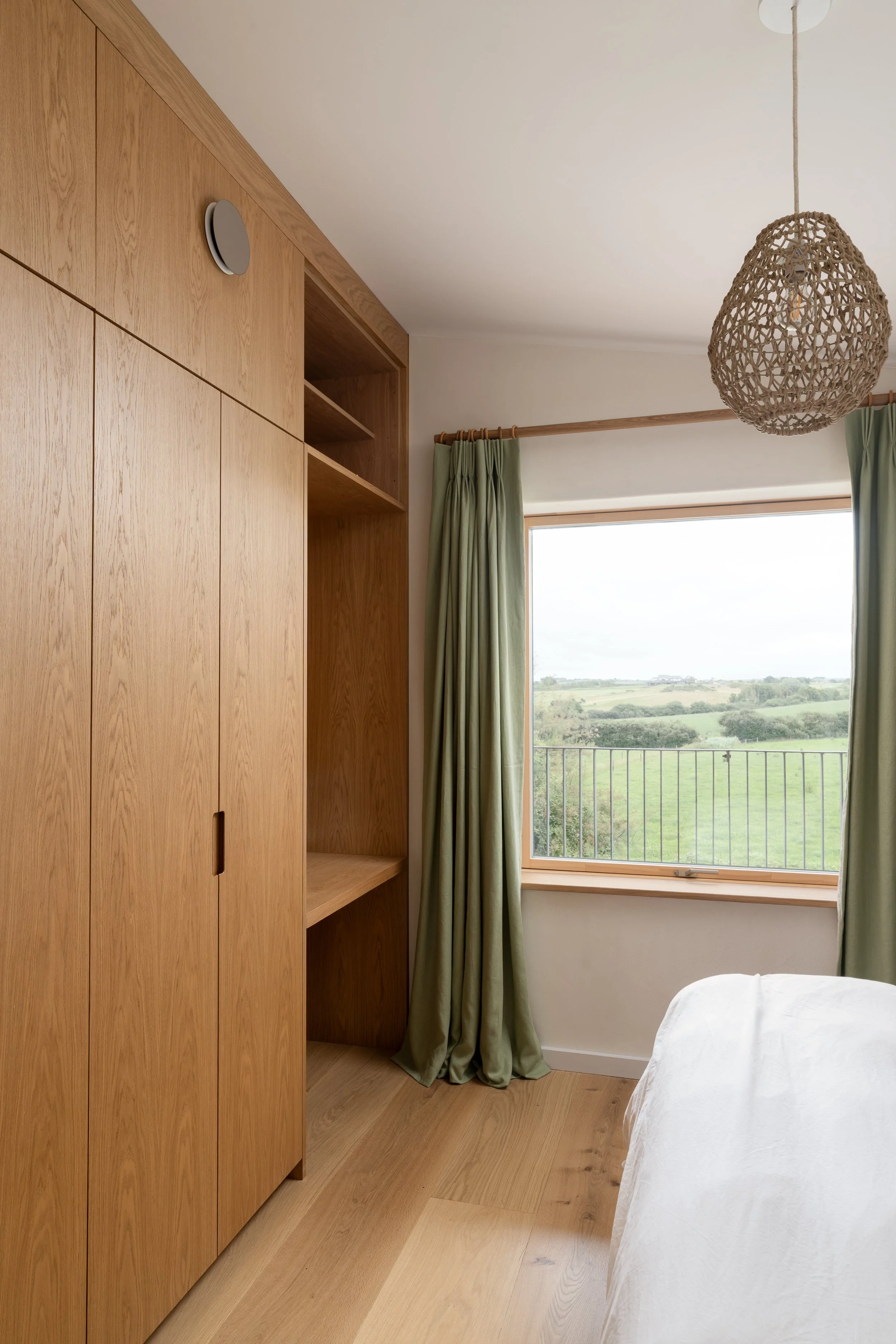 A bedroom with wooden cabinetry, green curtains, a large window with a view of a green landscape, and a woven light fixture hanging from the ceiling.
