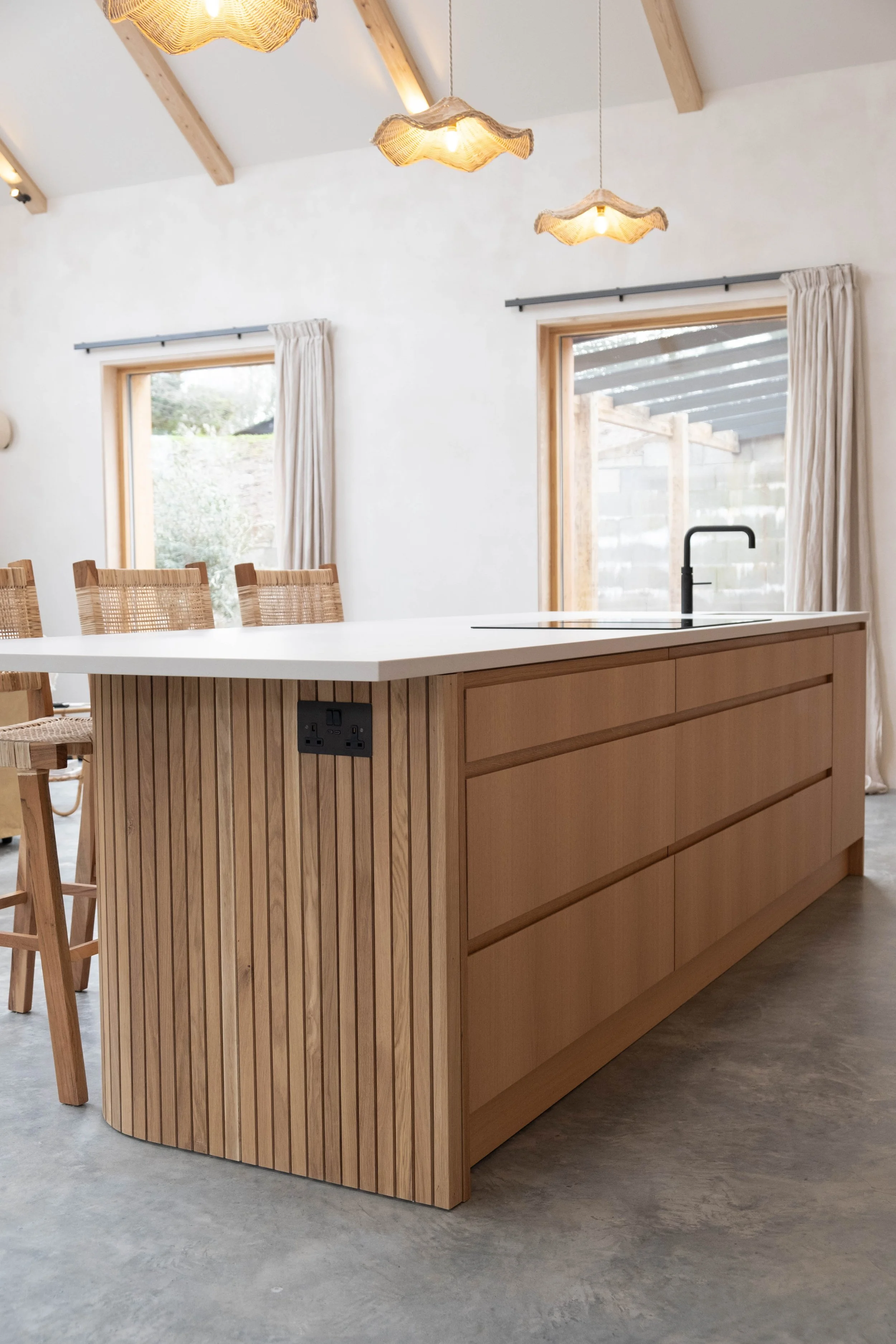 A wooden kitchen island with slatted sides, a white countertop, and a black faucet, situated in a modern kitchen with large windows and hanging pendant lights.