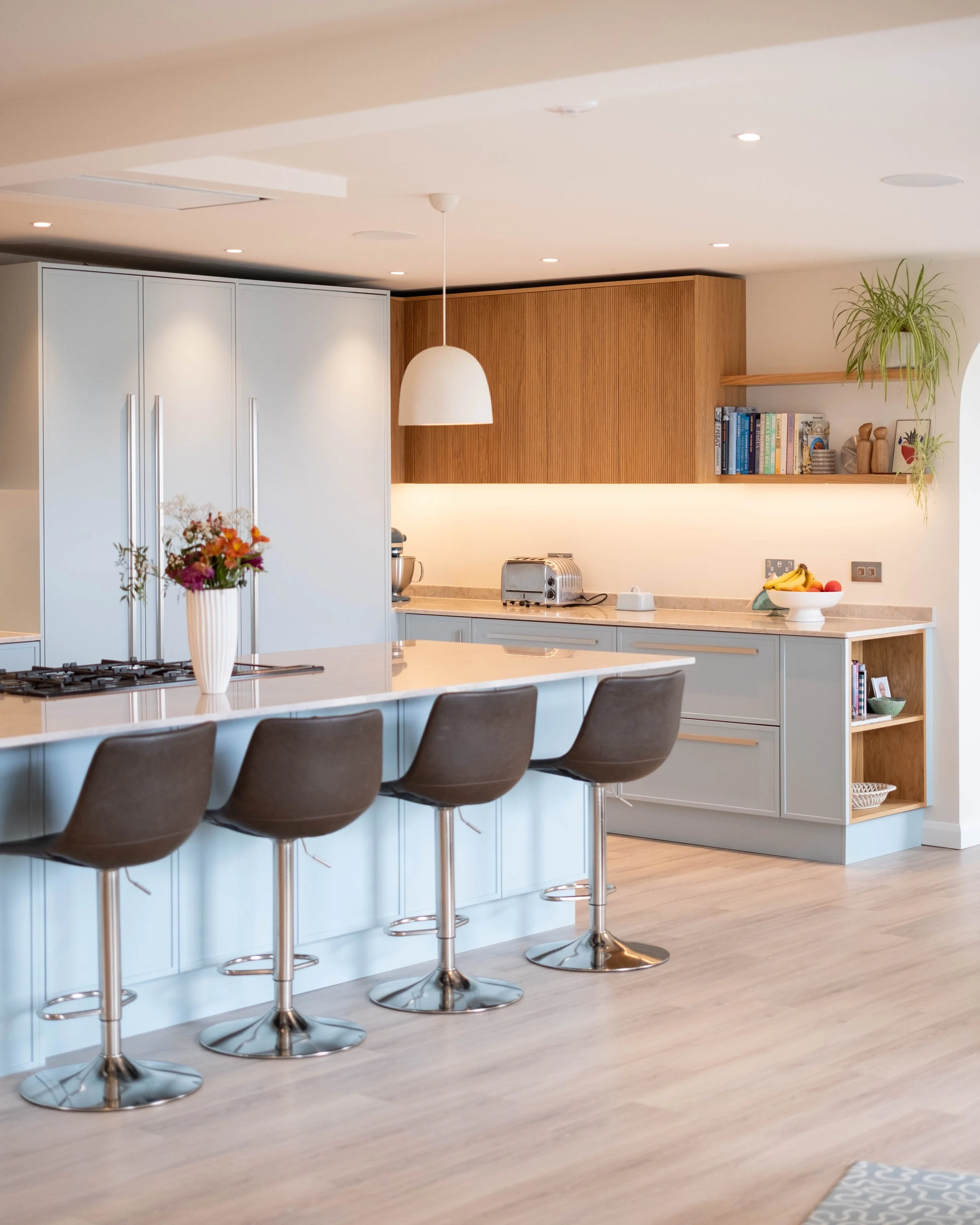 Modern kitchen with white cabinets, a marble island, black bar stools, open wooden shelves with books and plants, a hanging white pendant light, and a fruit bowl on the counter.