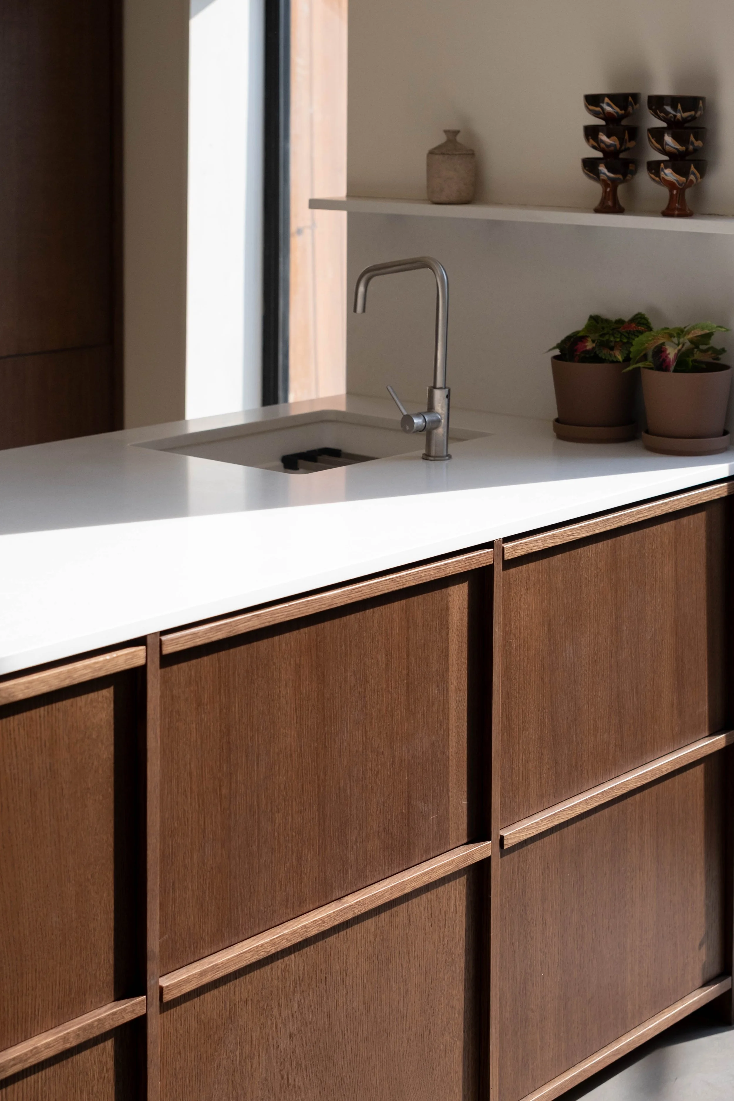 A kitchen with a white countertop, wooden cabinets, a modern silver faucet, and a shelf with decorative bowls, a small vase, and potted plants.