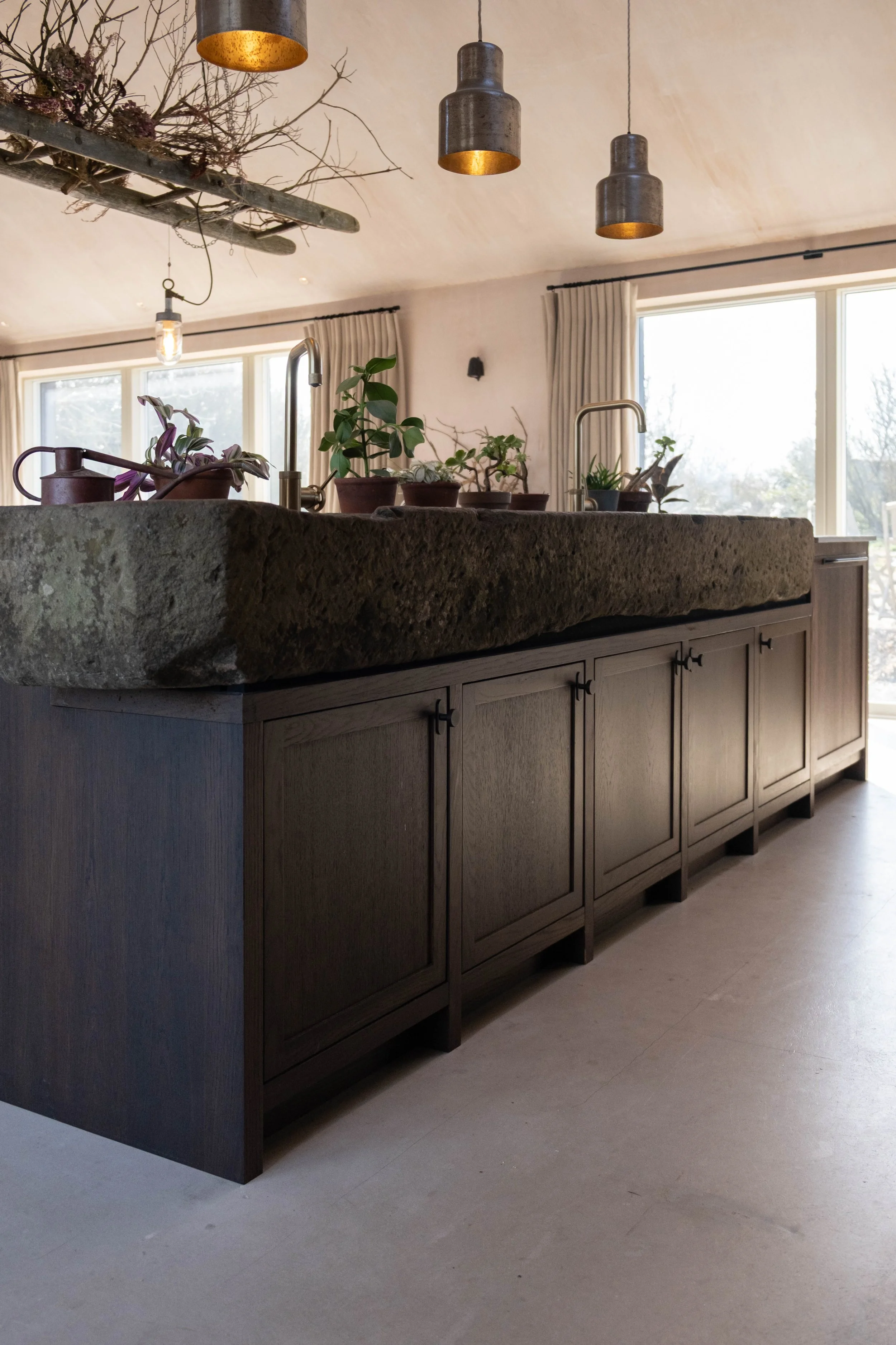 Modern kitchen with a large, rustic stone countertop, wooden cabinets, and potted plants on the surface. There are hanging pendant lights and large windows with curtains, letting in natural light.