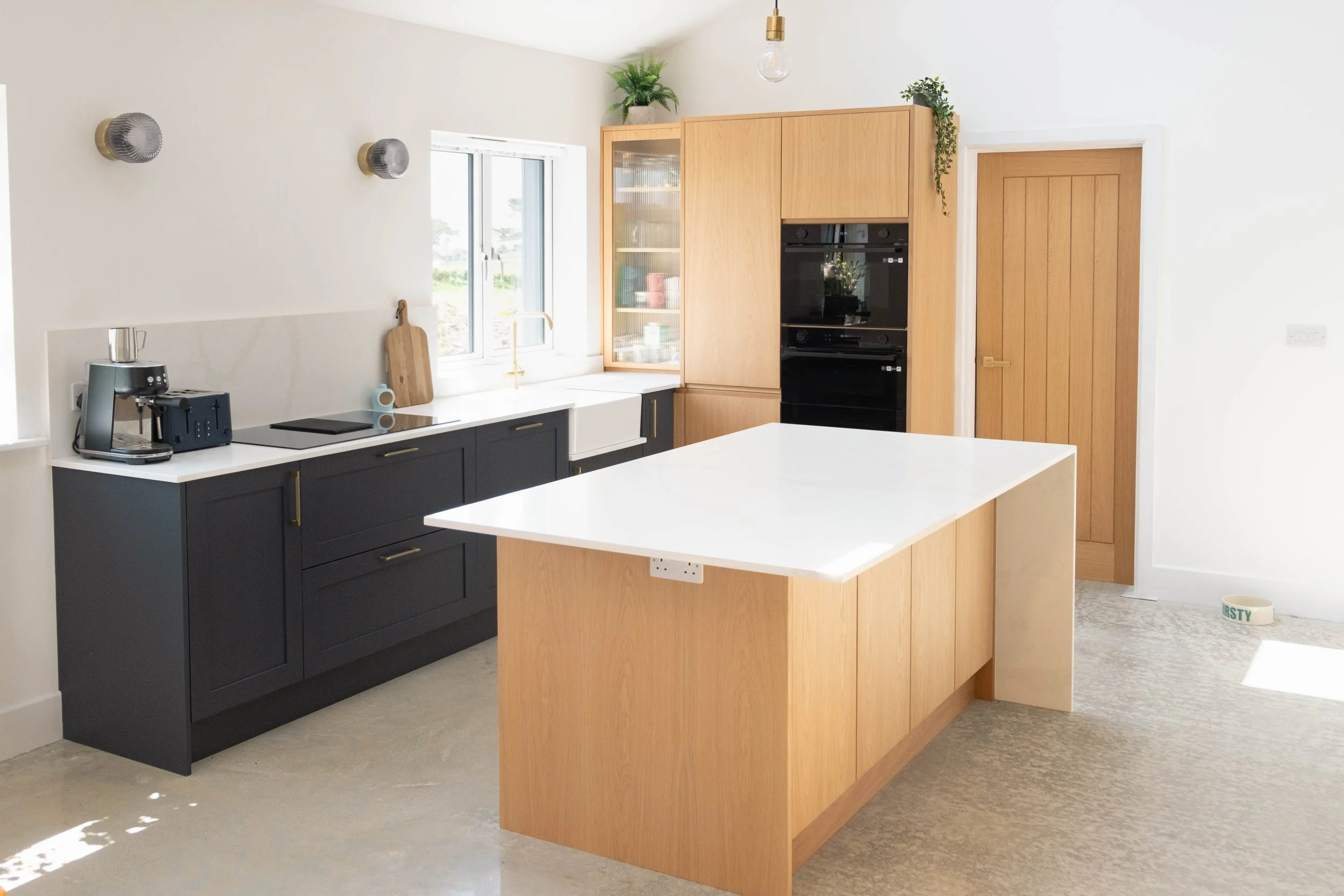 Modern kitchen with white upper walls, dark lower cabinets, and a wooden kitchen island. There is a window overlooking the outdoors, a built-in oven, a coffee machine, a cutting board, and some potted plants on a wooden cabinet.