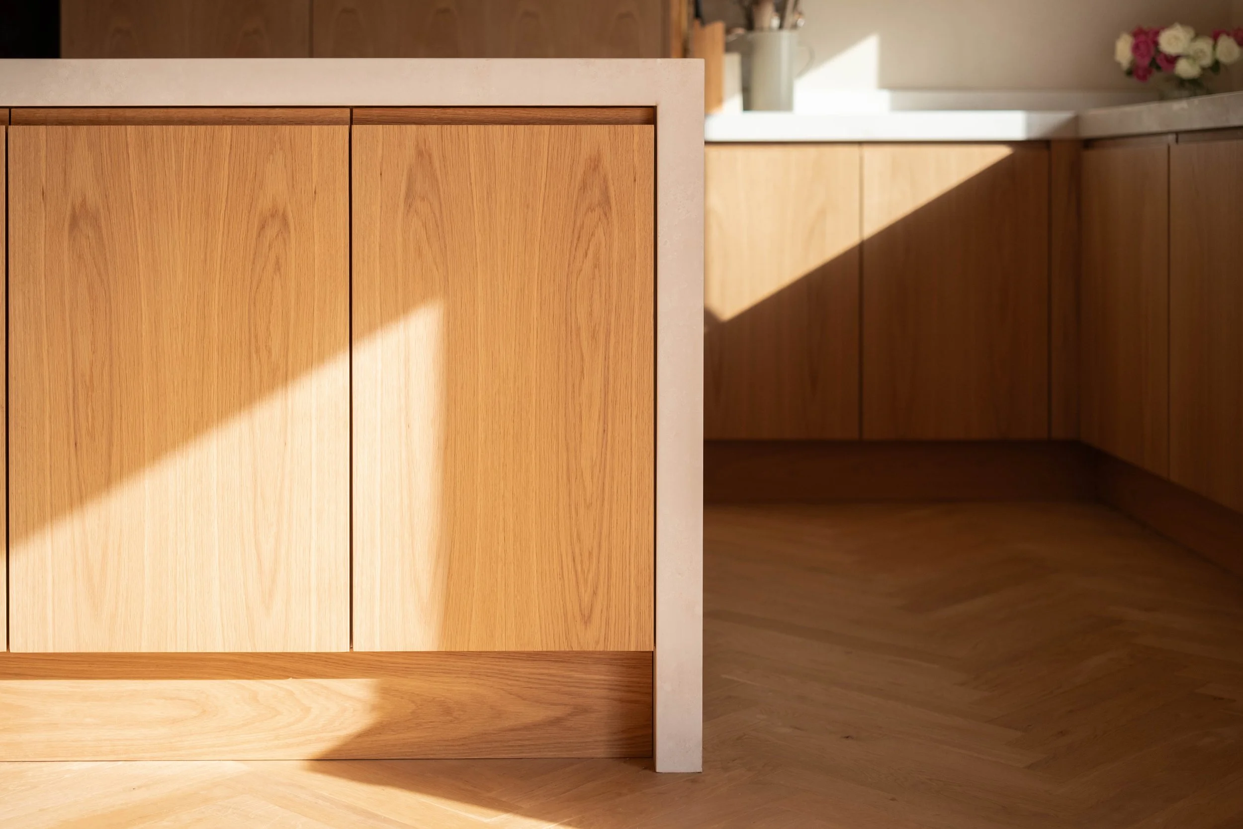 Close-up of a light wood kitchen cabinet with a white countertop, with sunlight casting a shadow across the floor and part of the cabinet.
