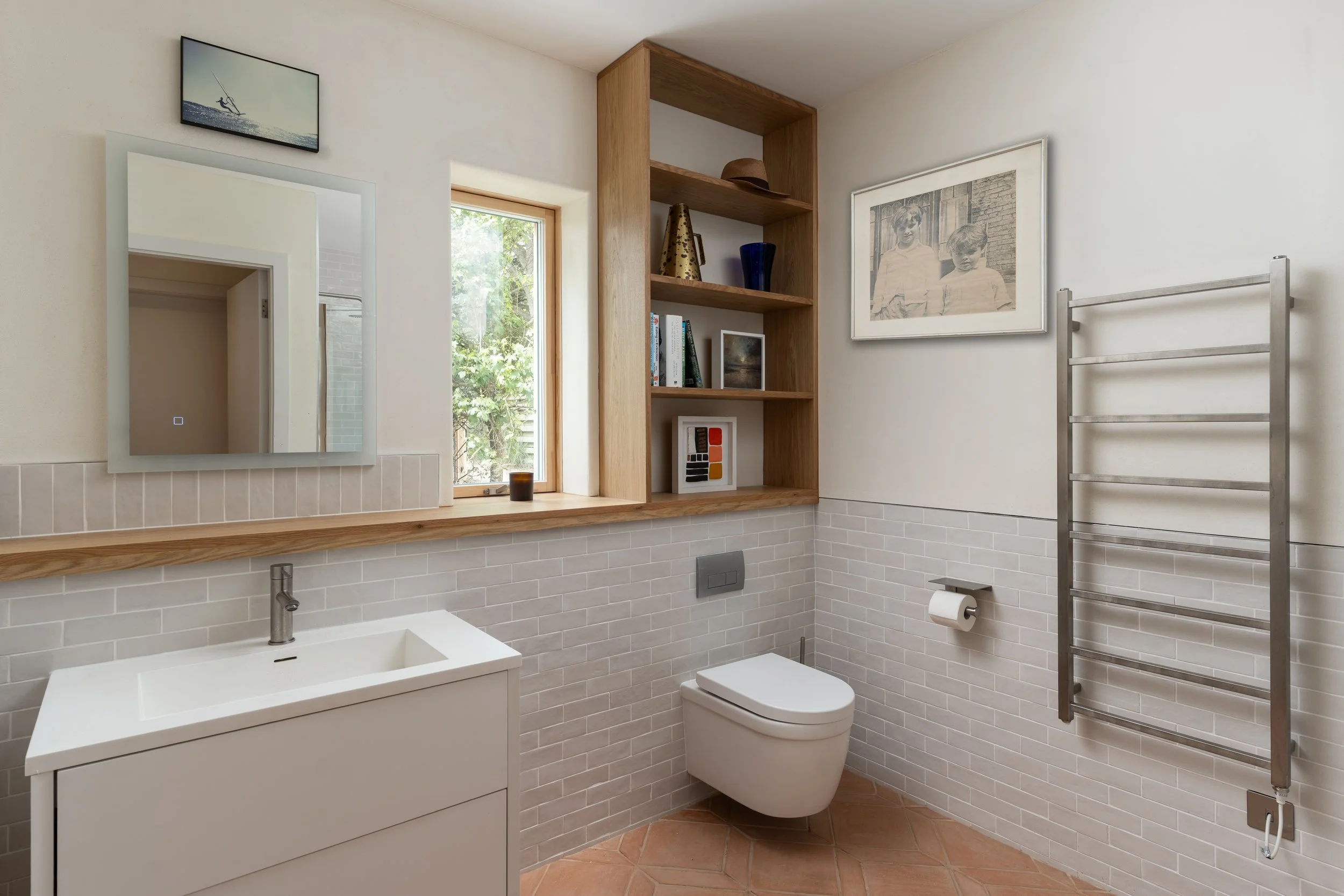 Modern bathroom with white subway tile walls, a small white sink with a chrome faucet, a toilet, a wooden shelf with books and decorative items, a framed black-and-white photo of two children, a ladder-style towel rack, and a small window with greene