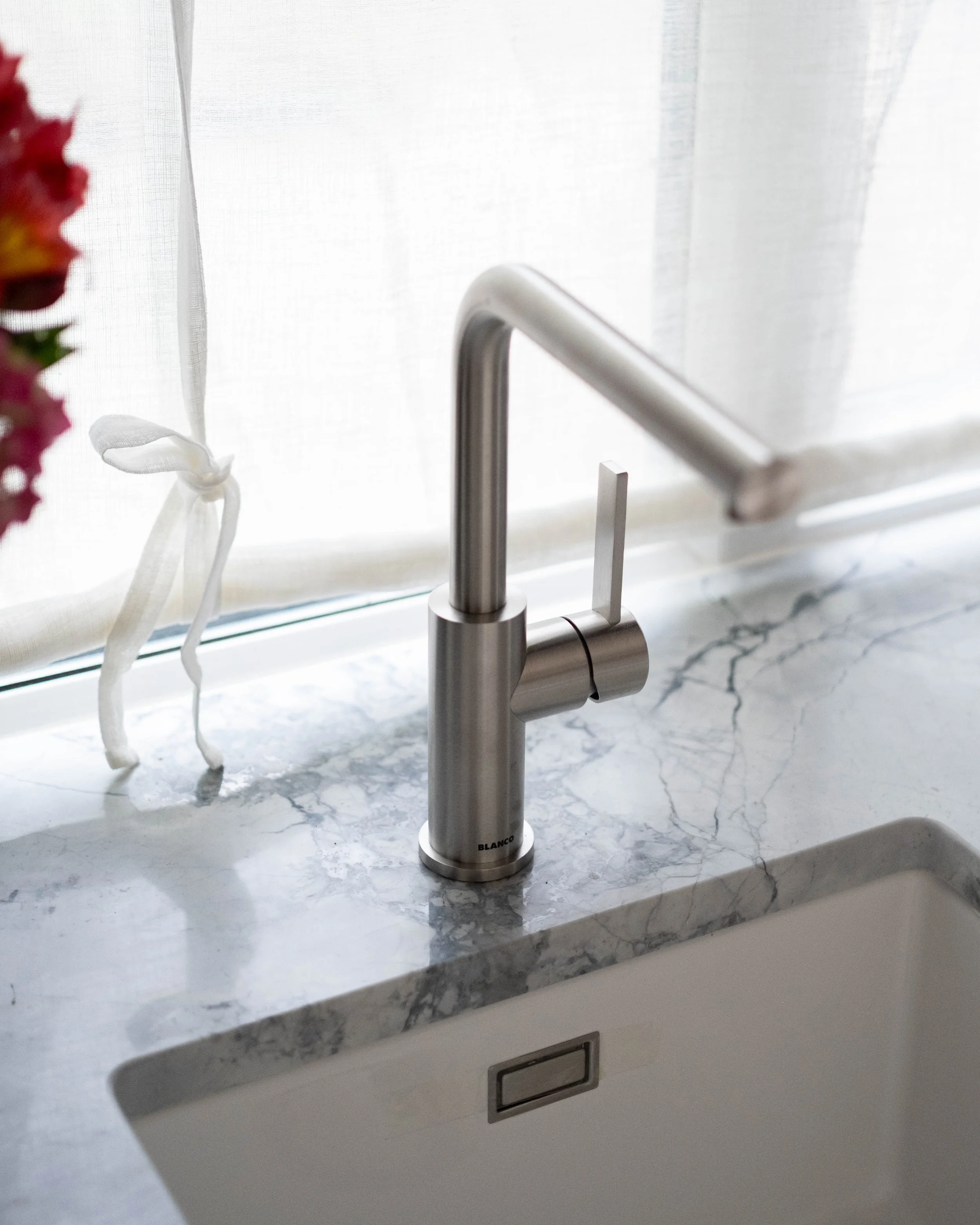 Close-up of a modern kitchen faucet on a marble countertop near a window with white curtains, partially visible.