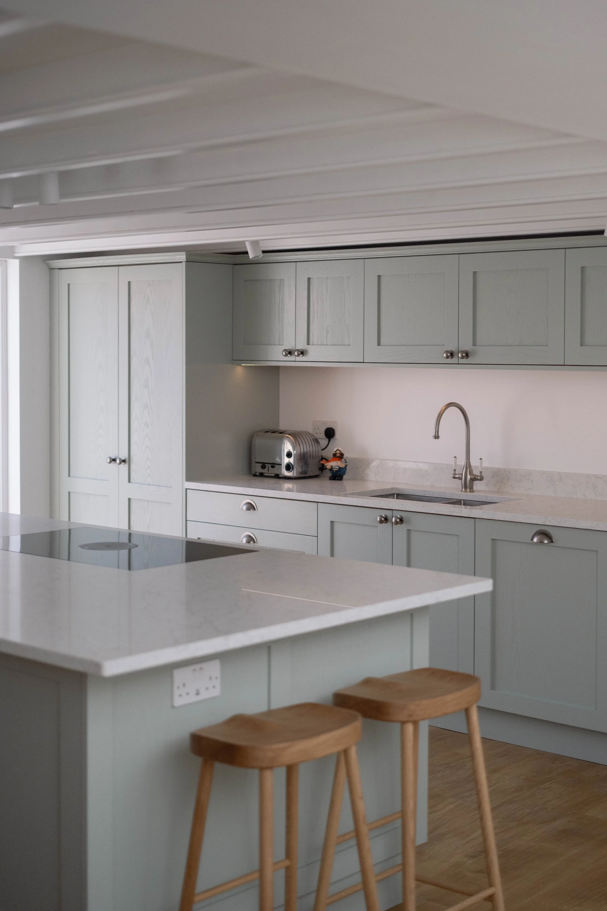 A modern kitchen with white cabinets, a marble countertop, a stainless steel sink, and a toaster on the counter. There are three wooden bar stools at a white kitchen island with electrical outlets.
