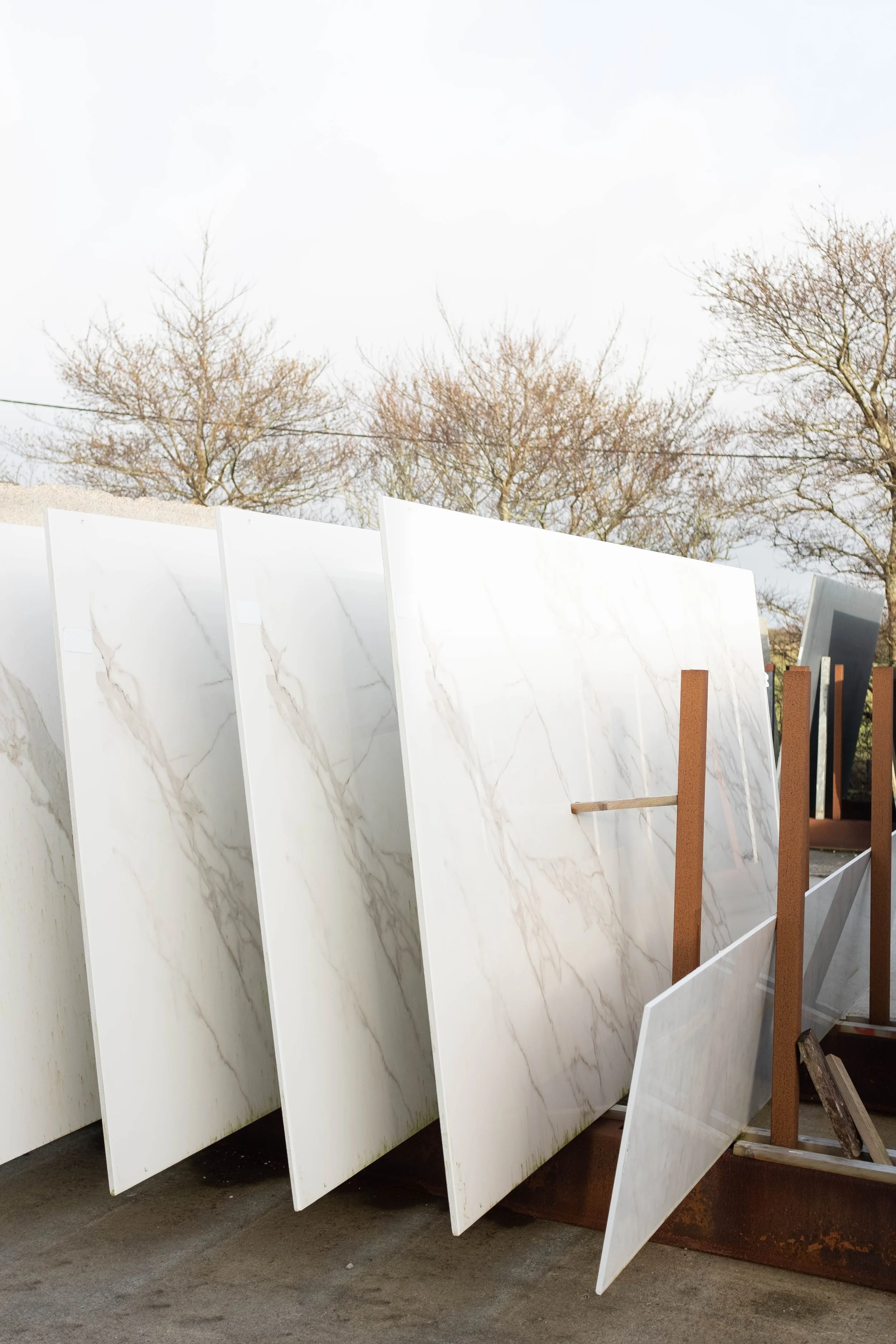 Large marble slabs leaning against wooden support beams outside on a concrete surface with leafless trees in the background.