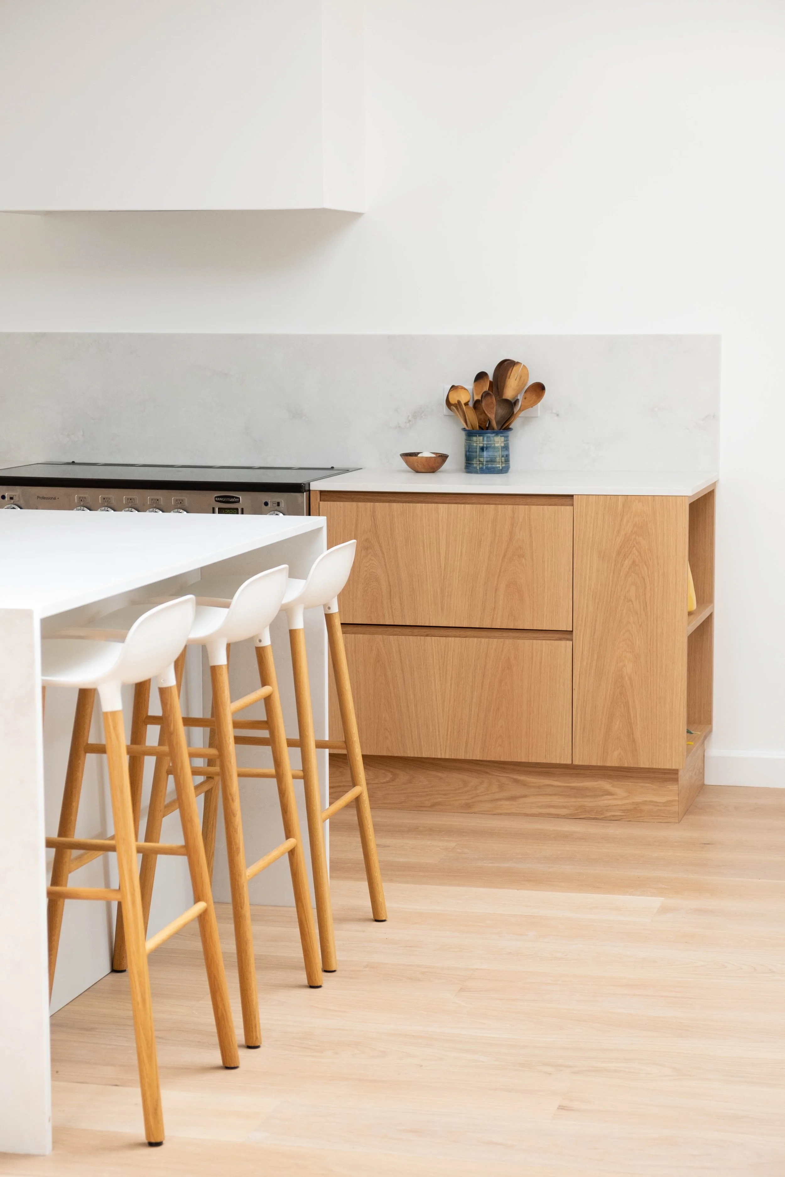 A kitchen with a white island counter, a wooden cabinet, a marble backsplash, and a blue ceramic jar holding wooden spoons. There are white chairs with wooden legs around the island, and light wood flooring.