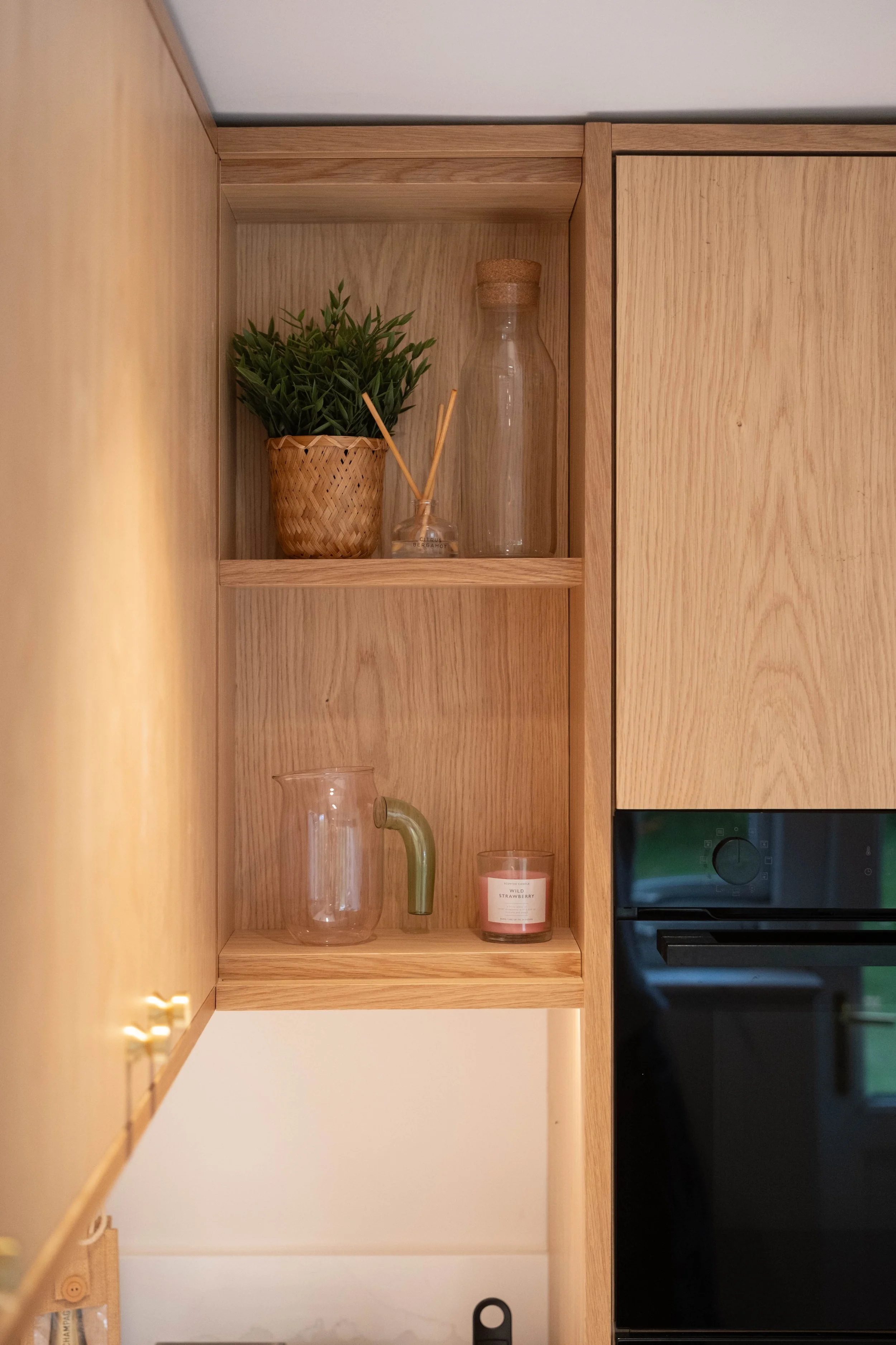 Wooden kitchen cabinet with decorative items including a potted green plant, a small reed diffuser, a glass bottle, a clear glass jug with a green handle, and a pink candle labeled 'wild strawberry' on a shelf.