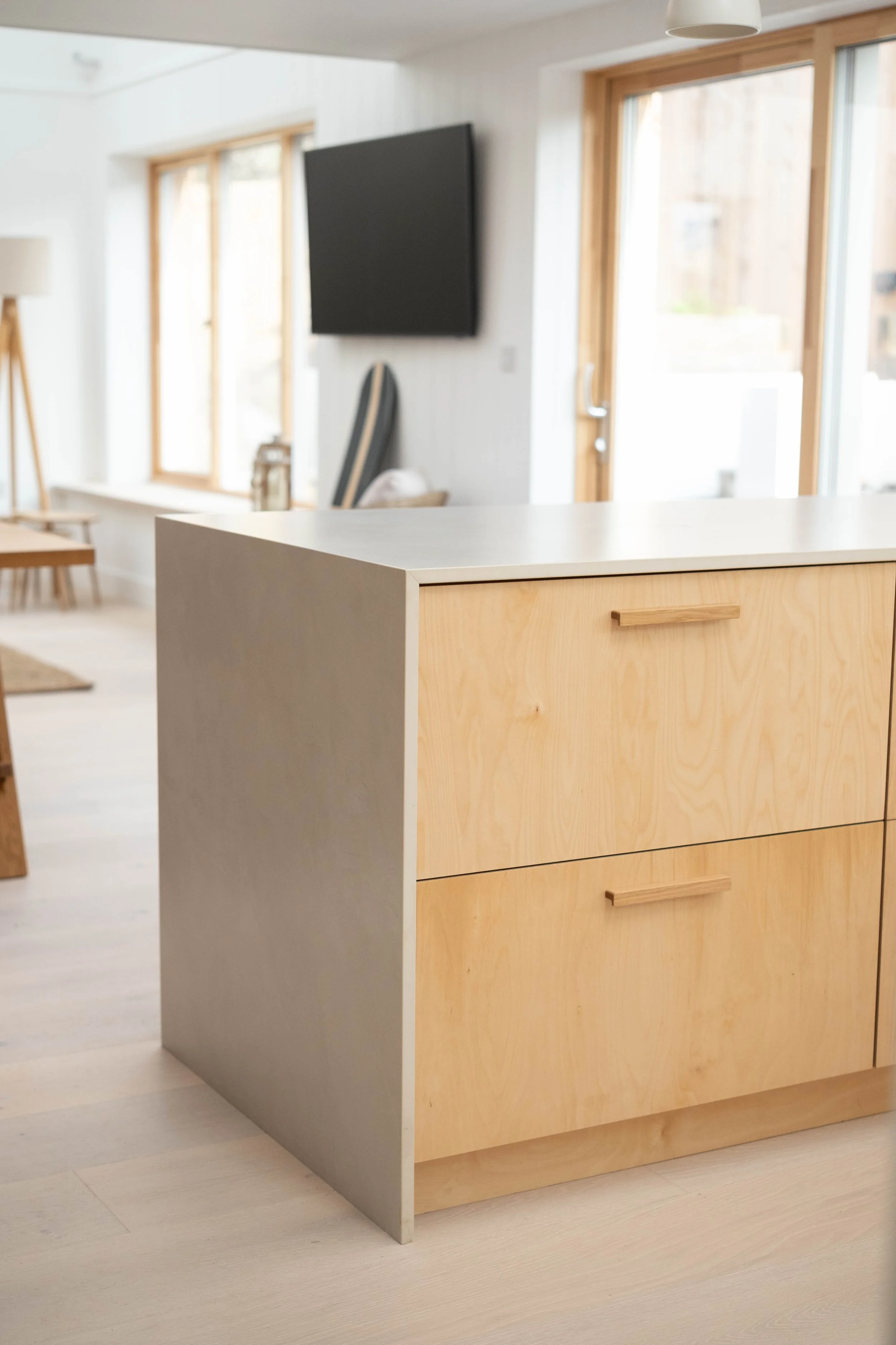 Modern kitchen island with wooden drawers and a white countertop, against large windows in a bright room.
