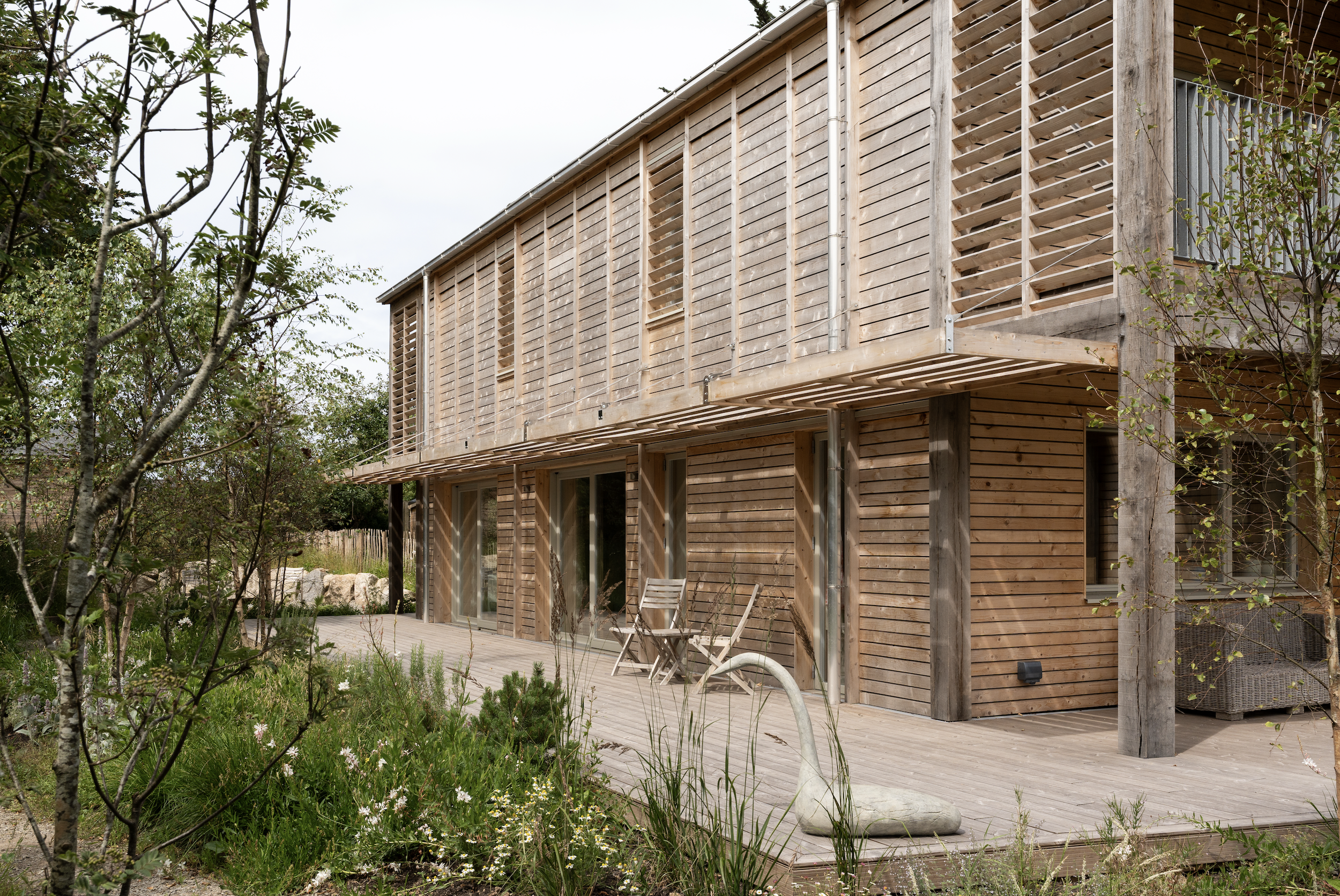 A modern wooden house with a balcony, outdoor patio, and garden surrounded by trees and plants.
