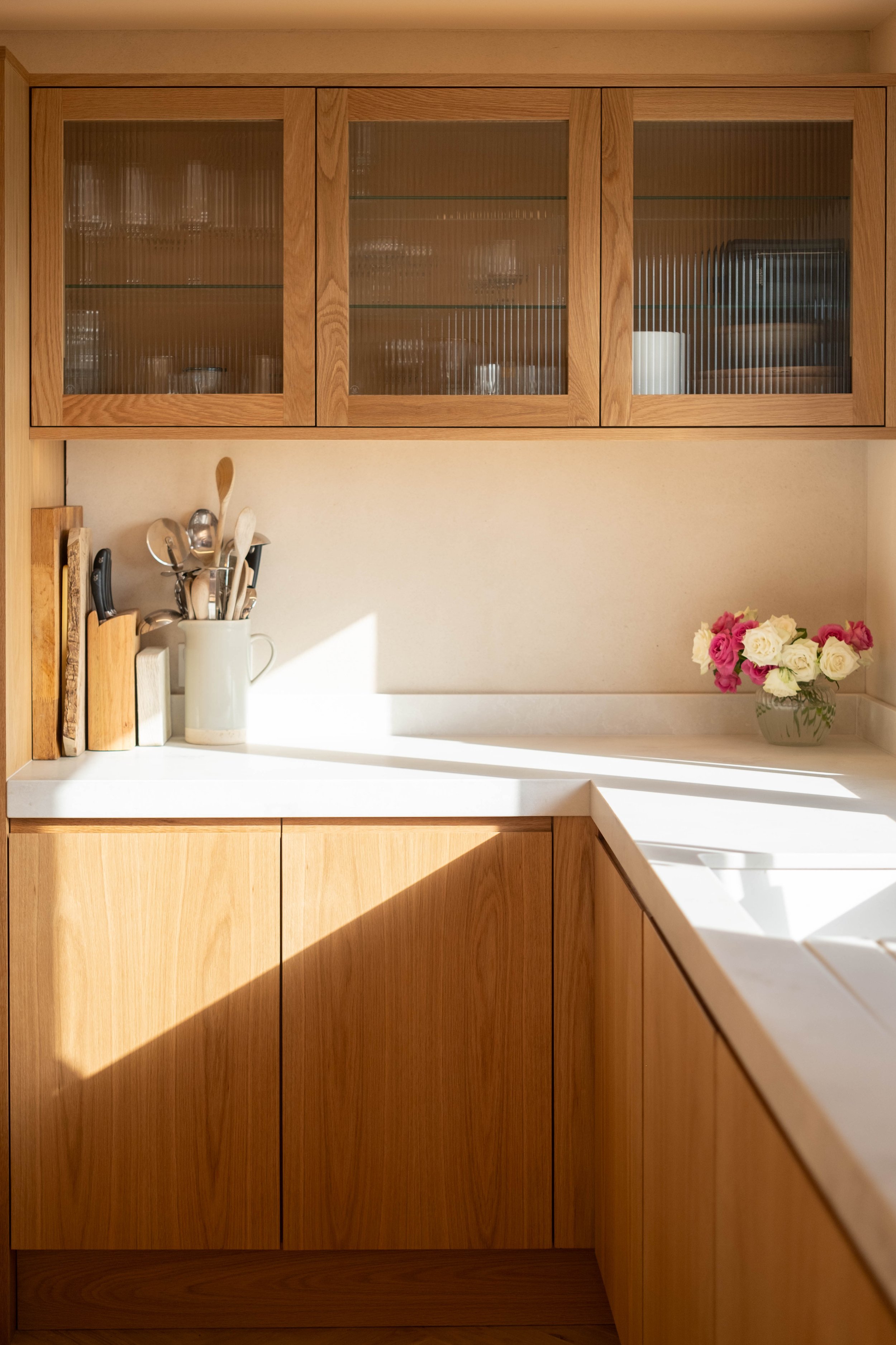 Kitchen with wooden cabinets, glass-front upper cabinets, a white countertop, a white utensil holder with silver utensils, some books, and a bouquet of pink and white roses on the counter.
