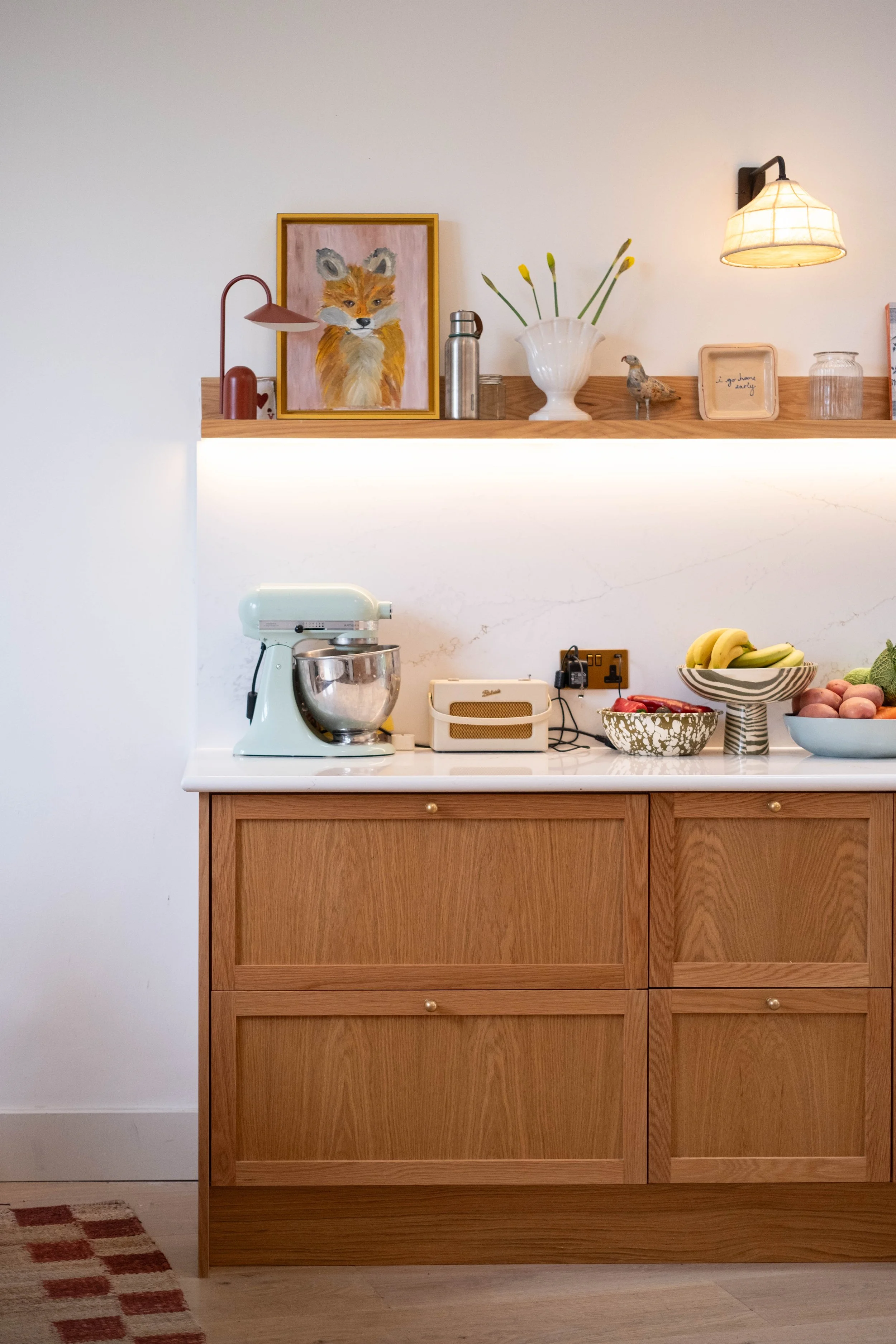 A kitchen countertop with a stand mixer, a fruit bowl containing bananas, and a small decorative bowl with strawberries. Above the countertop, there is a wooden shelf holding a framed painting of a fox, a vase with yellow flowers, a small bird figuri