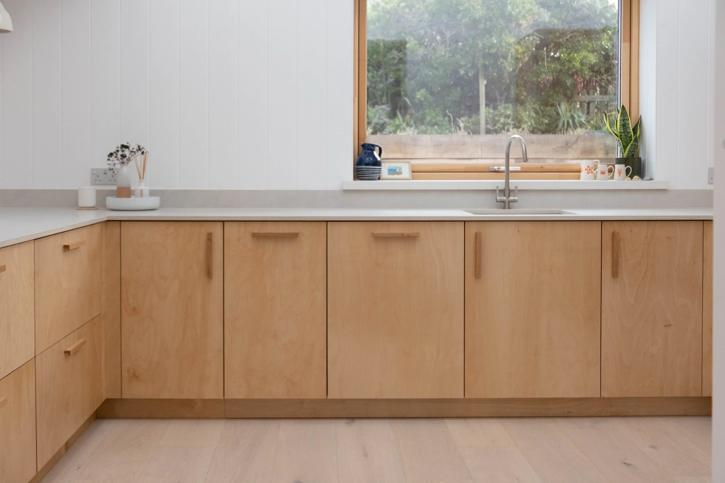 A modern kitchen with wooden cabinets, a white countertop, a window with a view of green trees outdoors, and decorative items including a plant, mugs, and a picture frame.