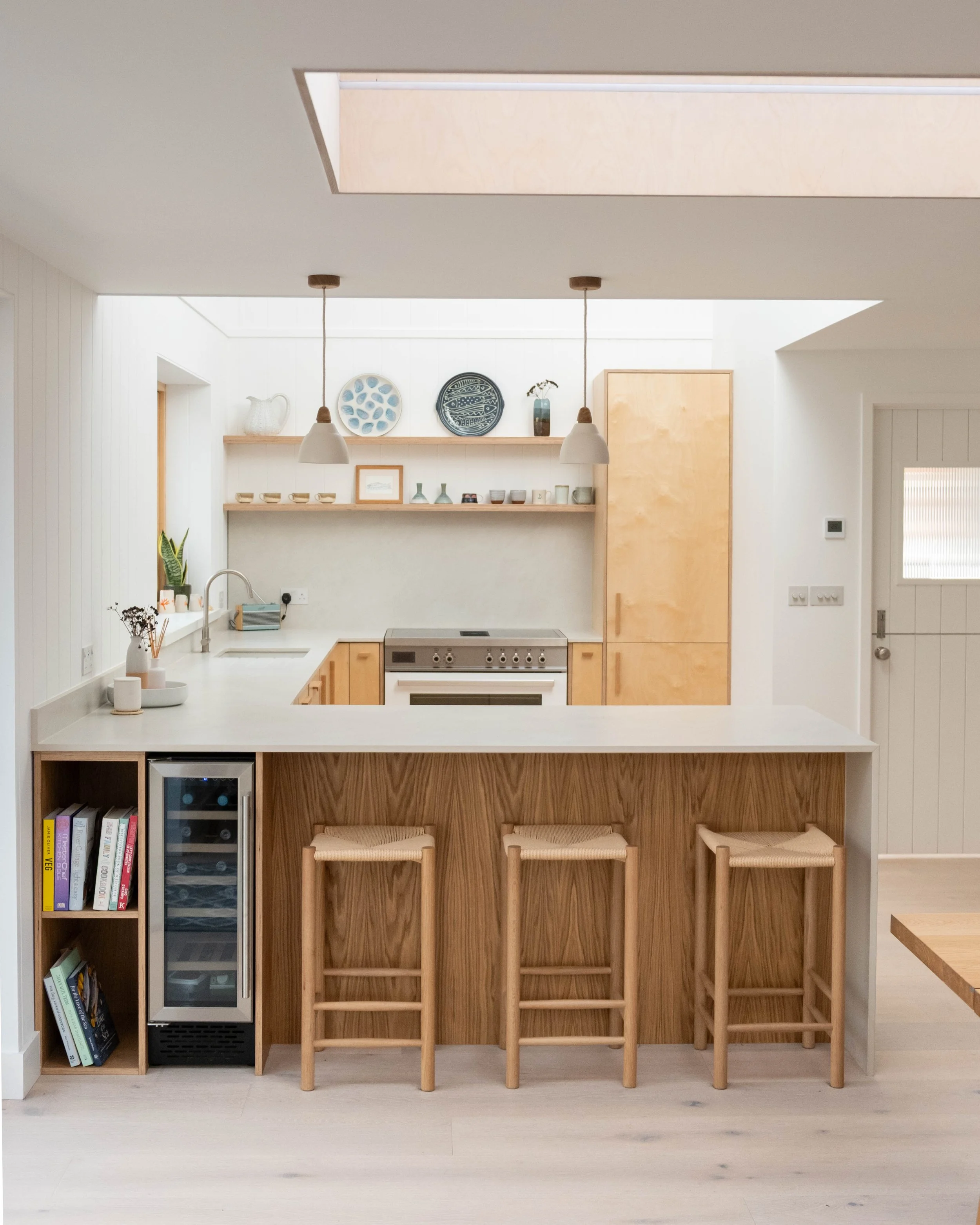 Modern minimalist kitchen with white walls, natural wood cabinets, and a white countertop. Three wooden stools with woven seats are at a breakfast bar. Open shelving displays decorative plates, vases, and bowls. Small wine fridge and books are on the